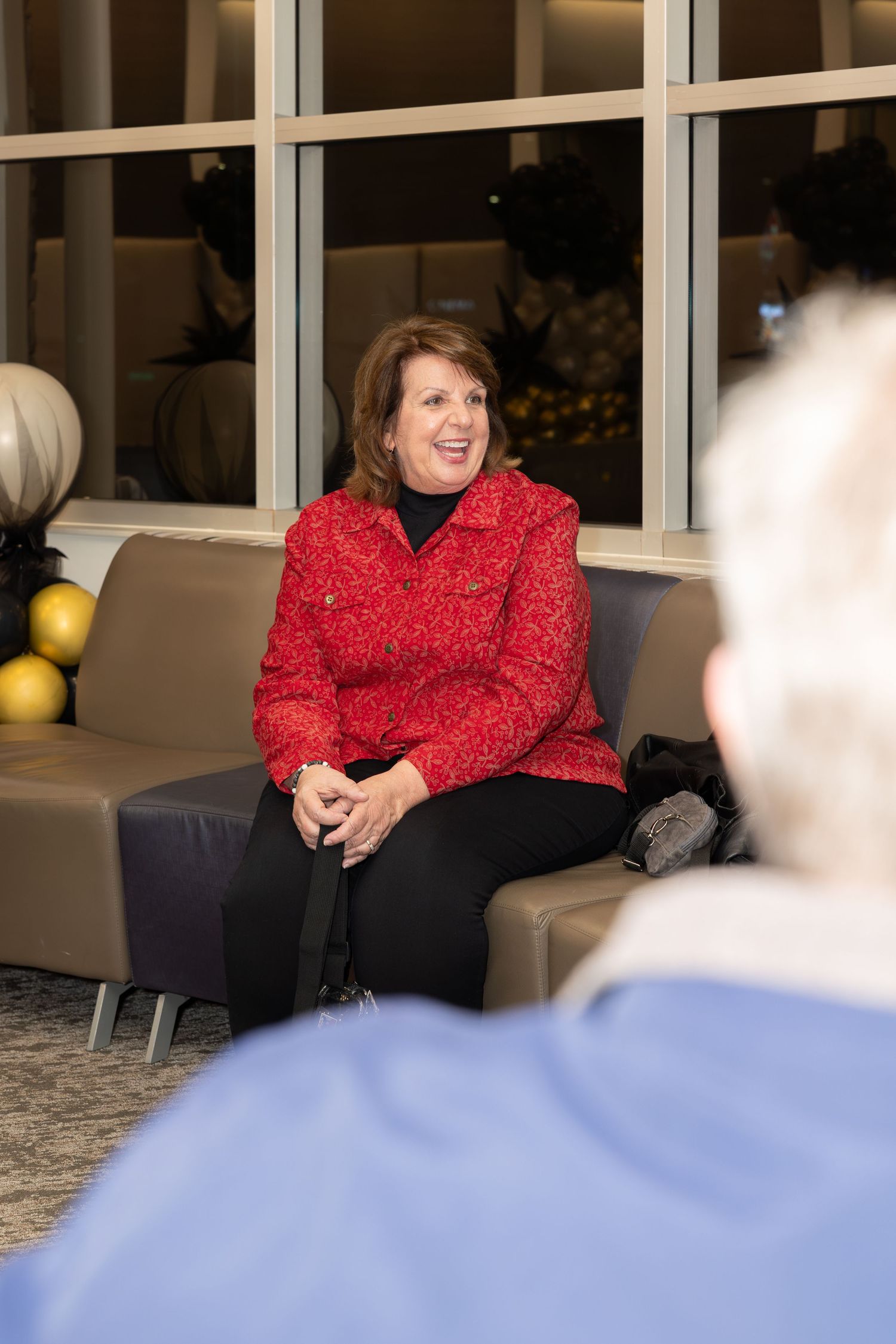 A person in a festive red sweater sits on a gray couch at an event with gold decorations in the background.
