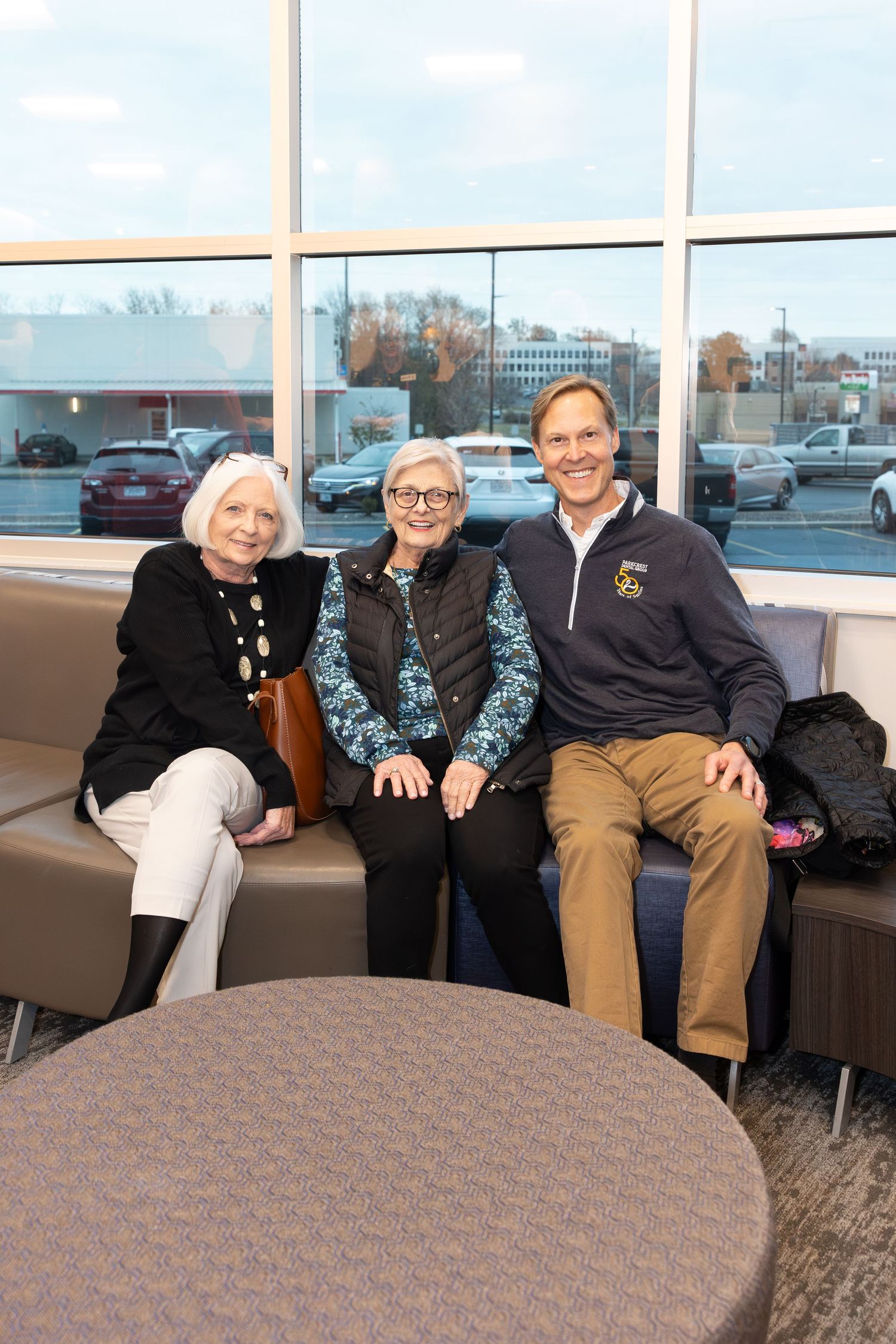 Three people sit together on a brown couch by large windows in an office setting.