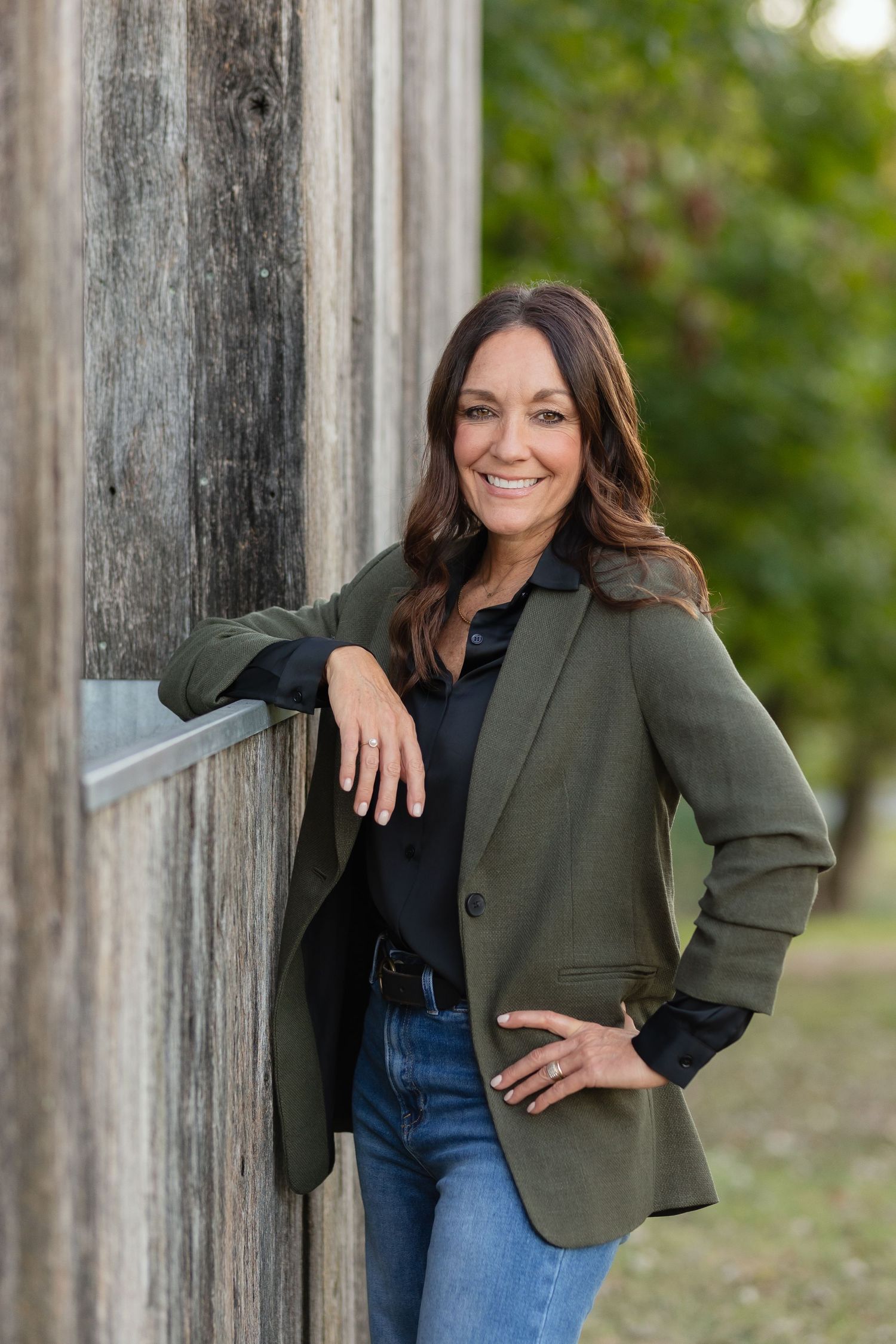 A professional headshot of someone in a green blazer and jeans leaning against a rustic wooden post.