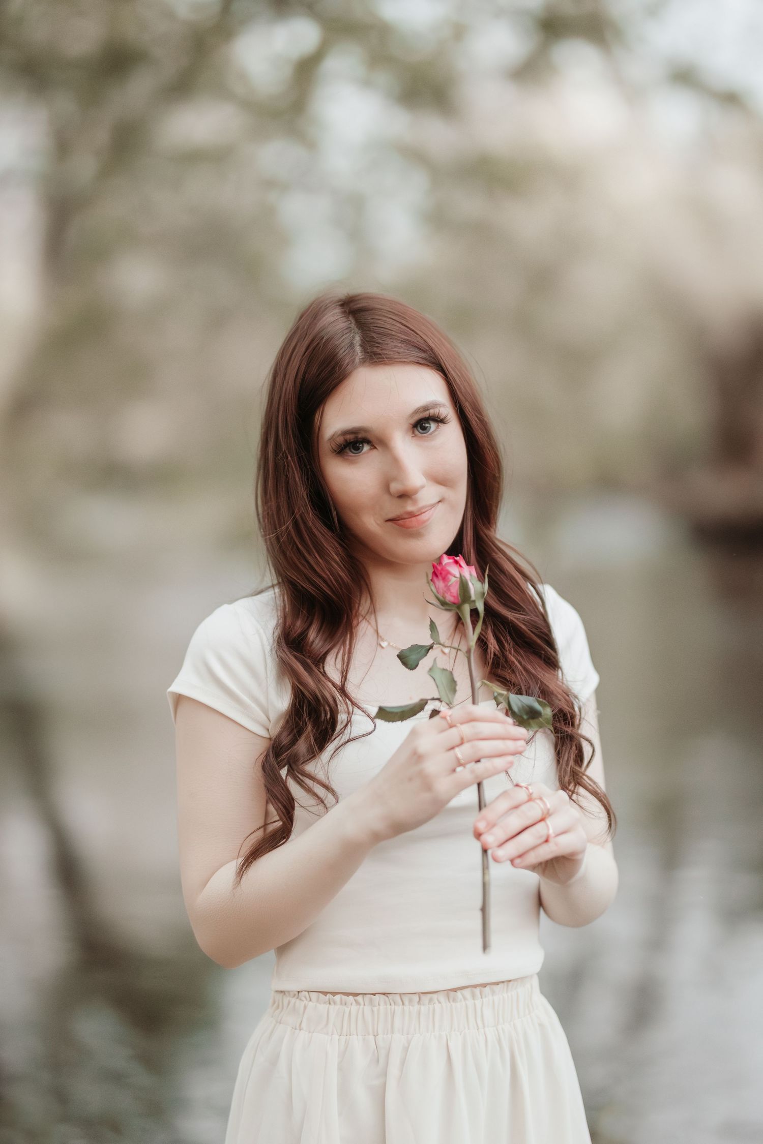 Woman in white dress holds pink rose in dreamy natural setting.