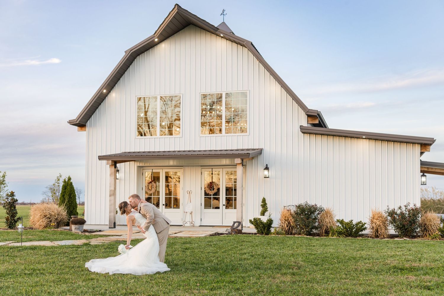 Elegant white barn wedding venue with couple dancing on the lawn at sunset.