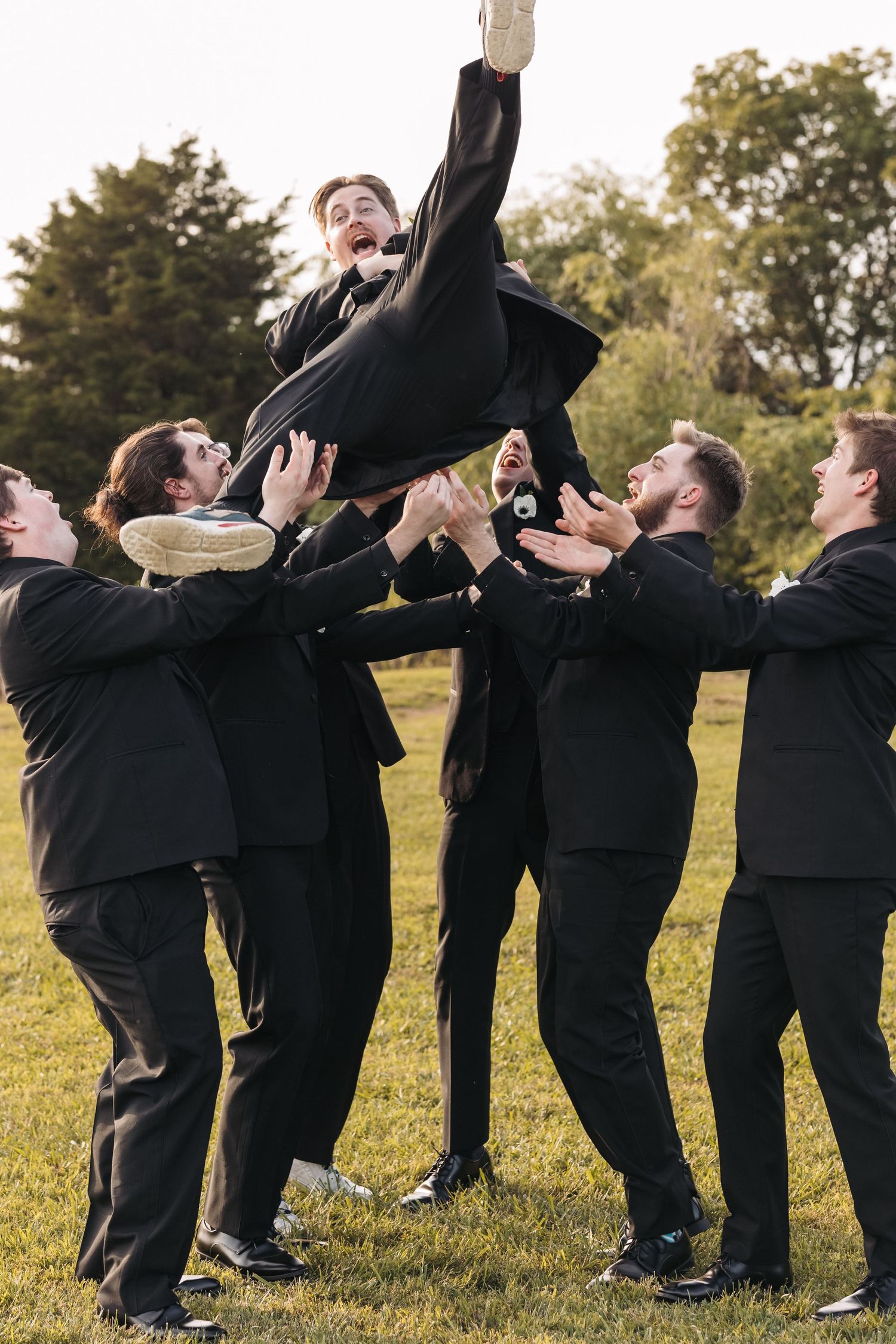 Groomsmen in black suits toss someone in the air during outdoor wedding photo session.