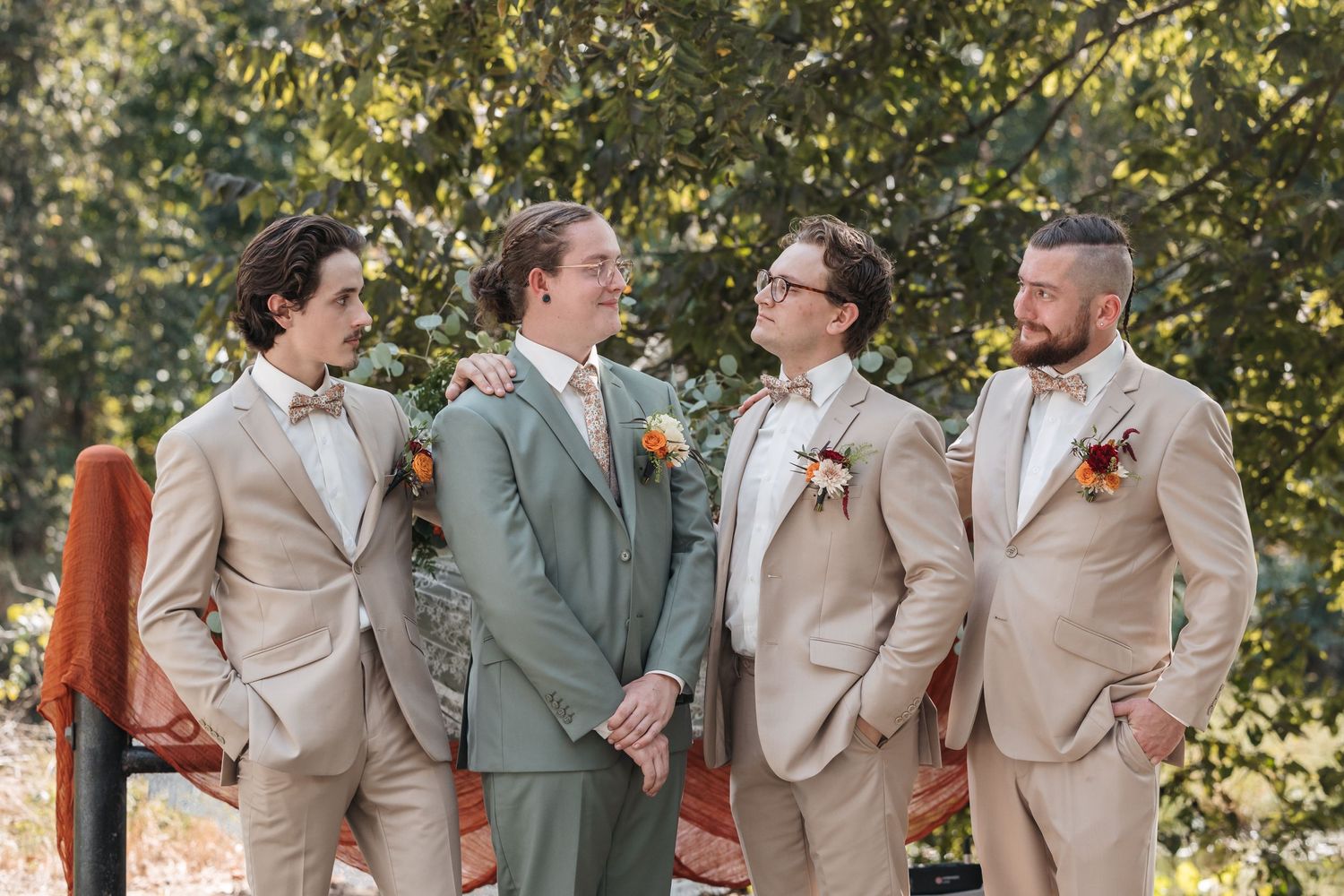 Groomsmen wearing light beige and sage green suits pose together at an outdoor wedding.
