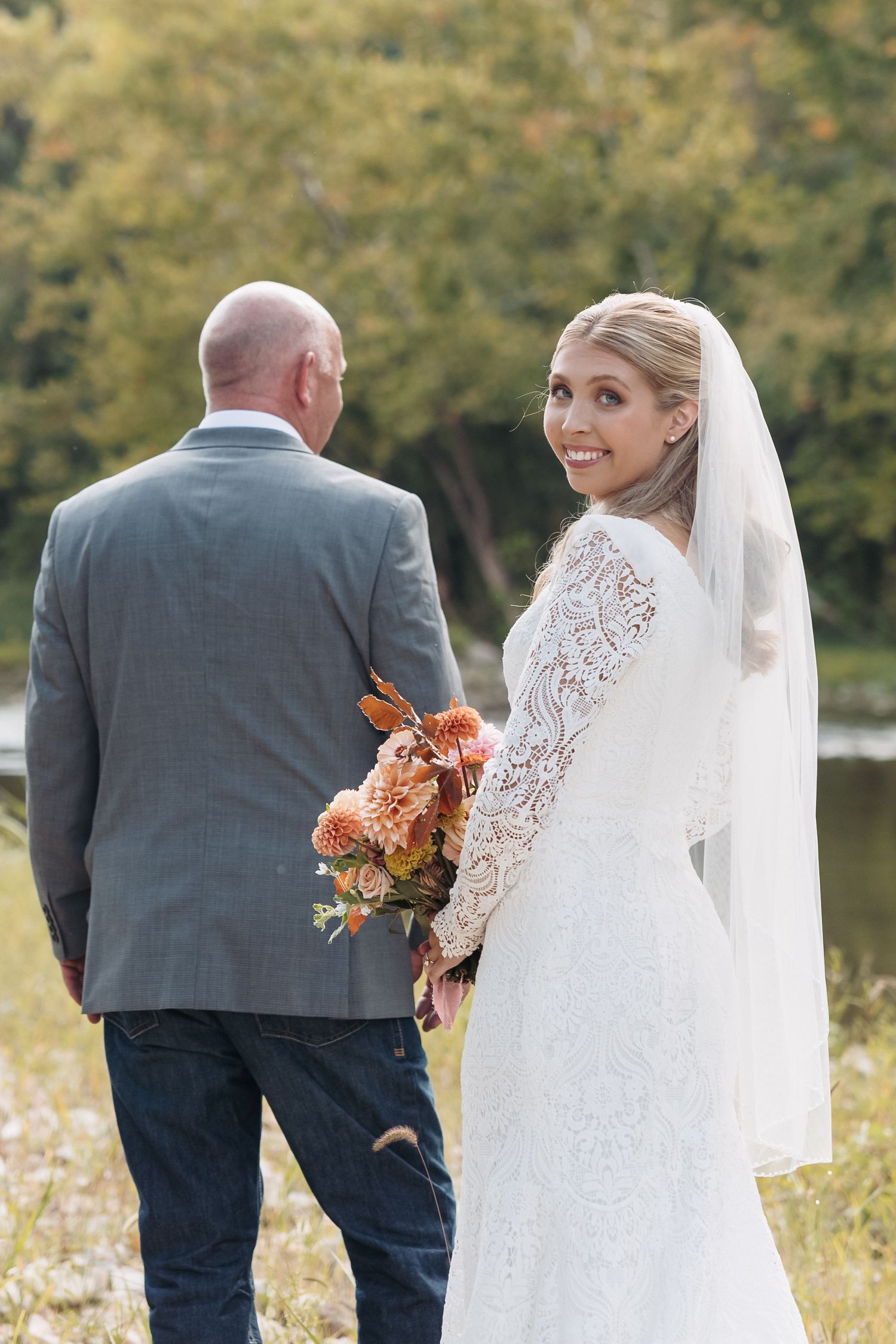 A couple in wedding attire poses with autumn-colored flowers by a scenic pond.