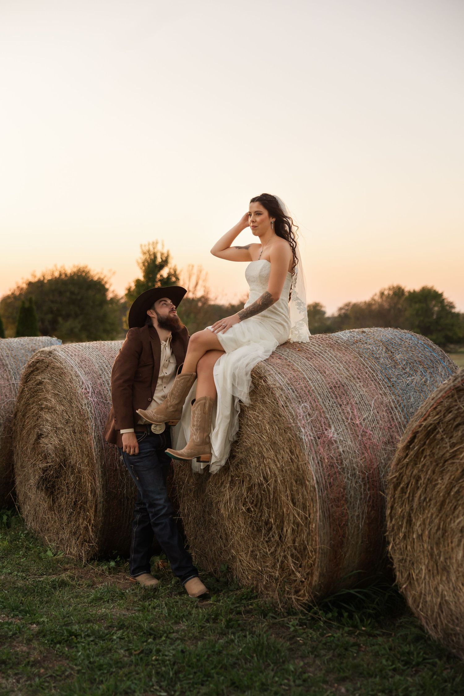 Couple in western attire at sunset, woman in white dress sitting on hay bale, man in cowboy hat leaning against it.