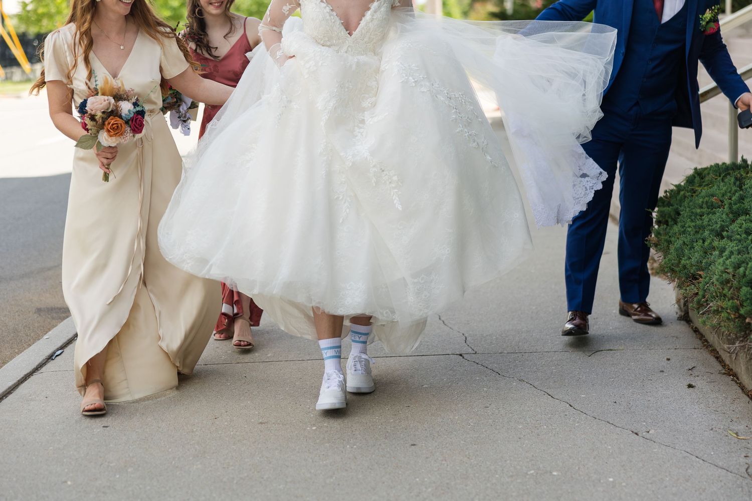 Bride in white ball gown walks in sneakers assisted by bridesmaids in gold and red dresses.