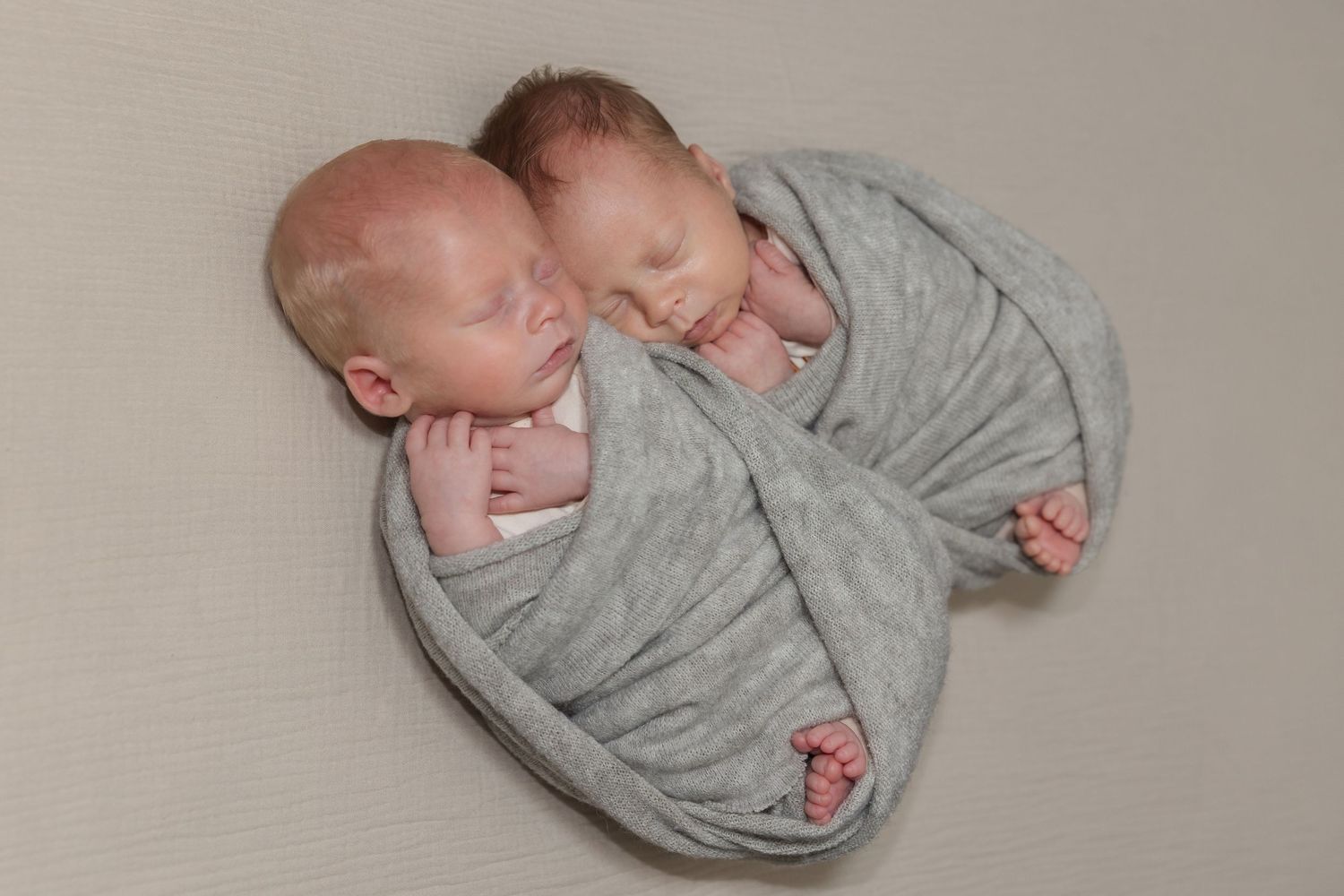 Two sleeping newborn twins swaddled together in a soft gray blanket on a neutral background.