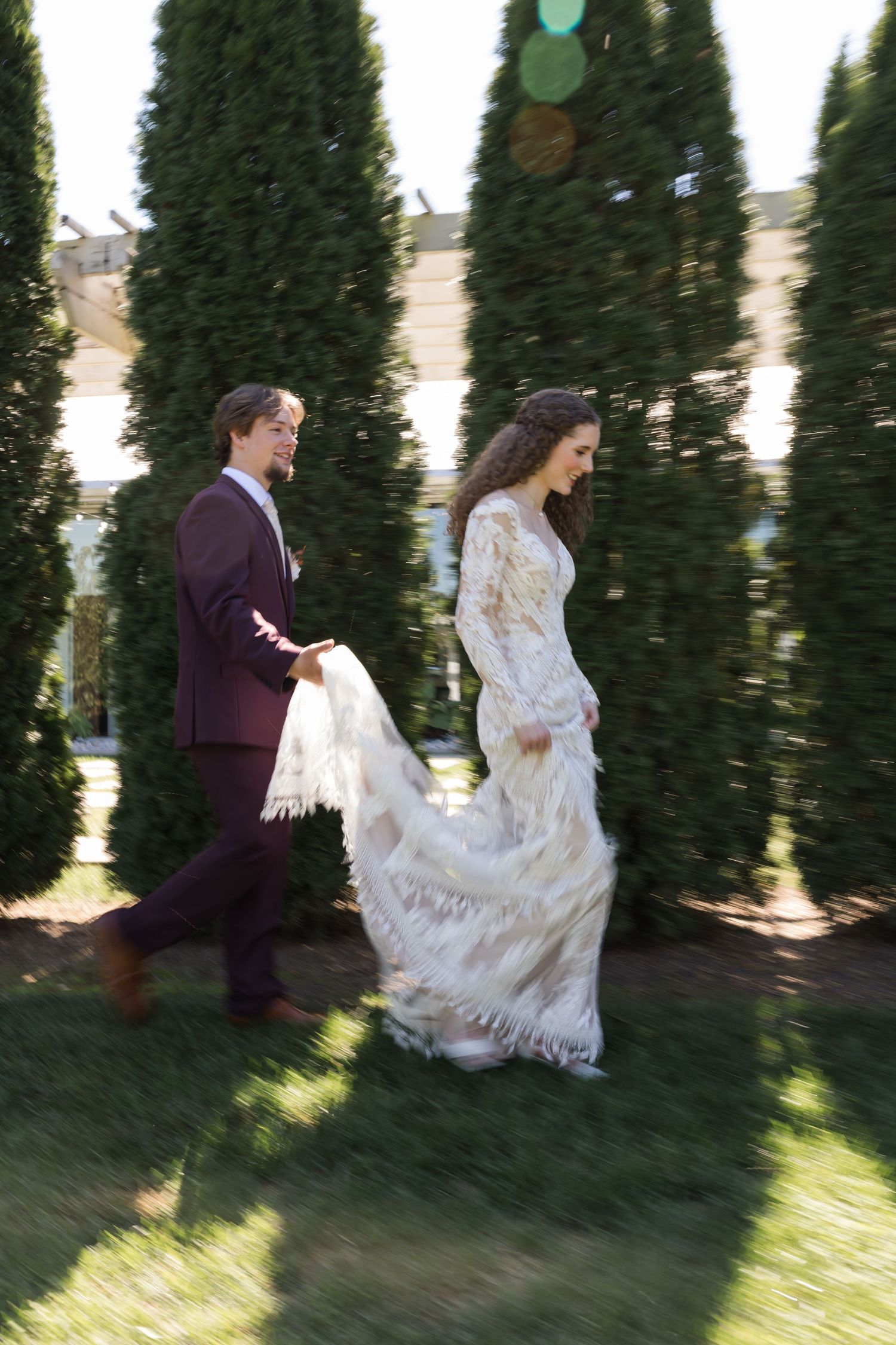 A couple in formal attire runs playfully through a garden of evergreen trees on a sunny afternoon.