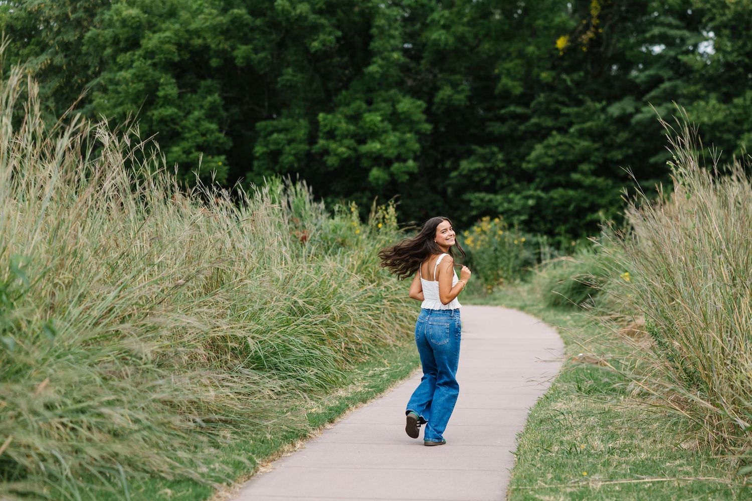 A figure in a floral dress walks along a scenic nature path surrounded by tall grass and green trees in summer.