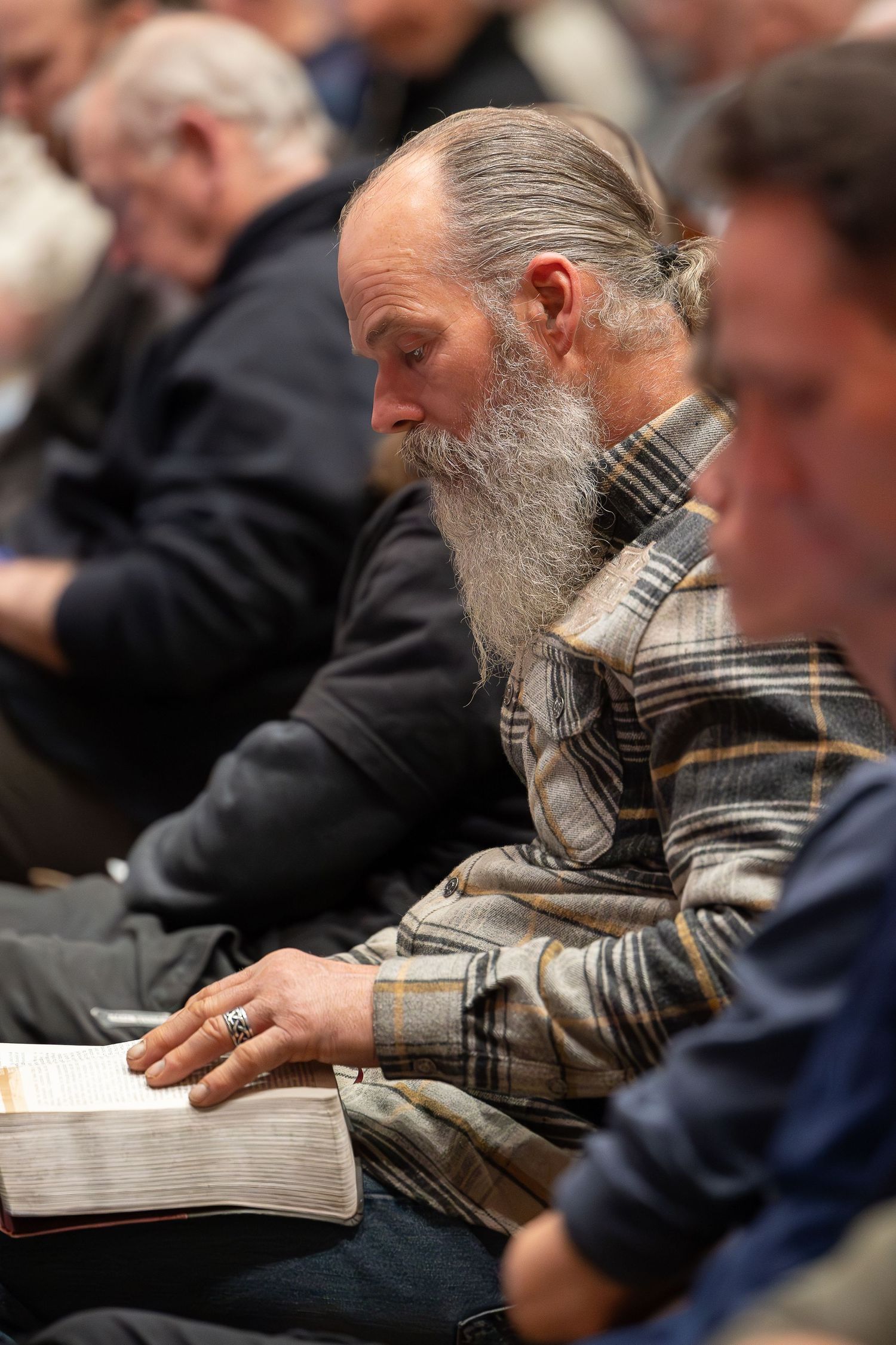 A person with a long gray beard sits in a meeting reading from an open book in a plaid shirt.