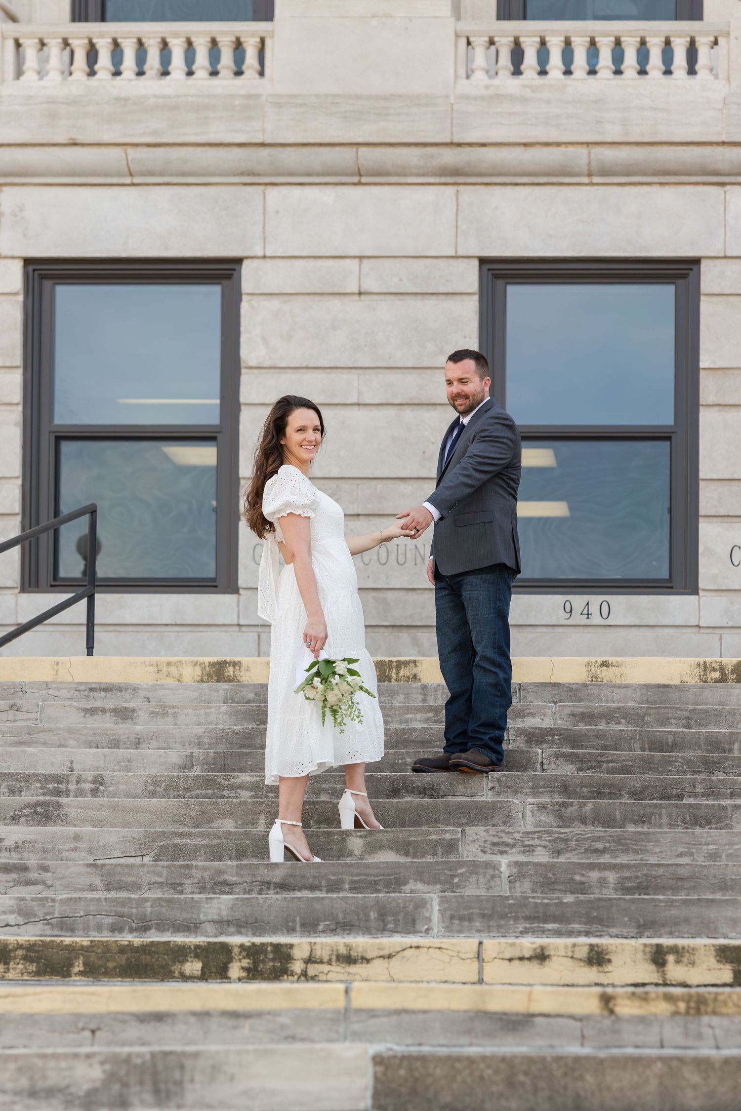 A couple stands on concrete steps outside a historic building with white stone walls and large windows.