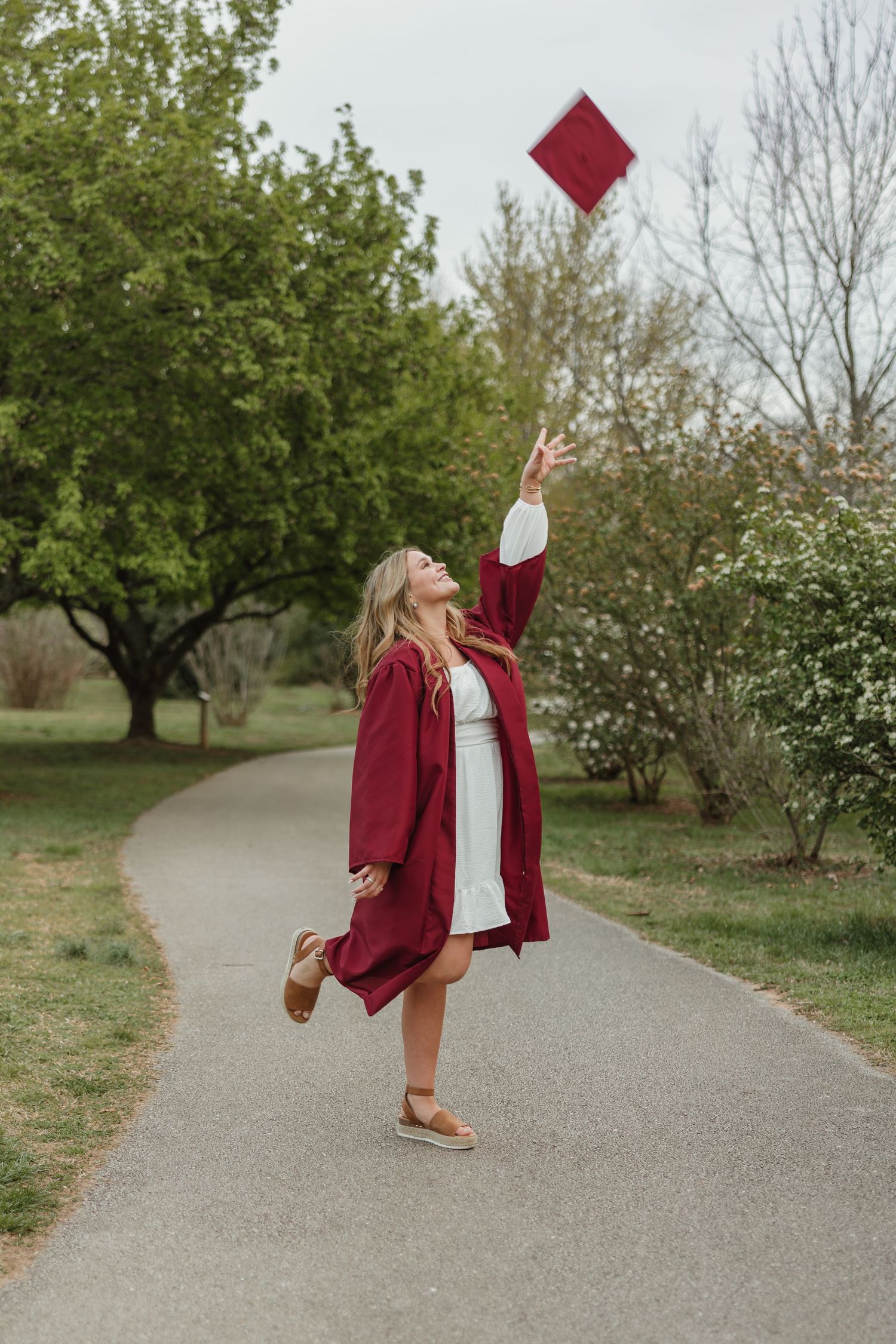Graduate in red cap and gown celebrates on a path by tossing their graduation cap in the air on a spring day.