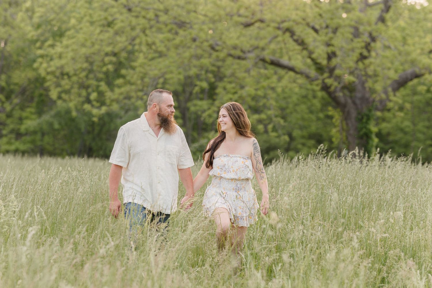 A couple walks hand in hand through a sunlit meadow of tall grass on a warm spring day.