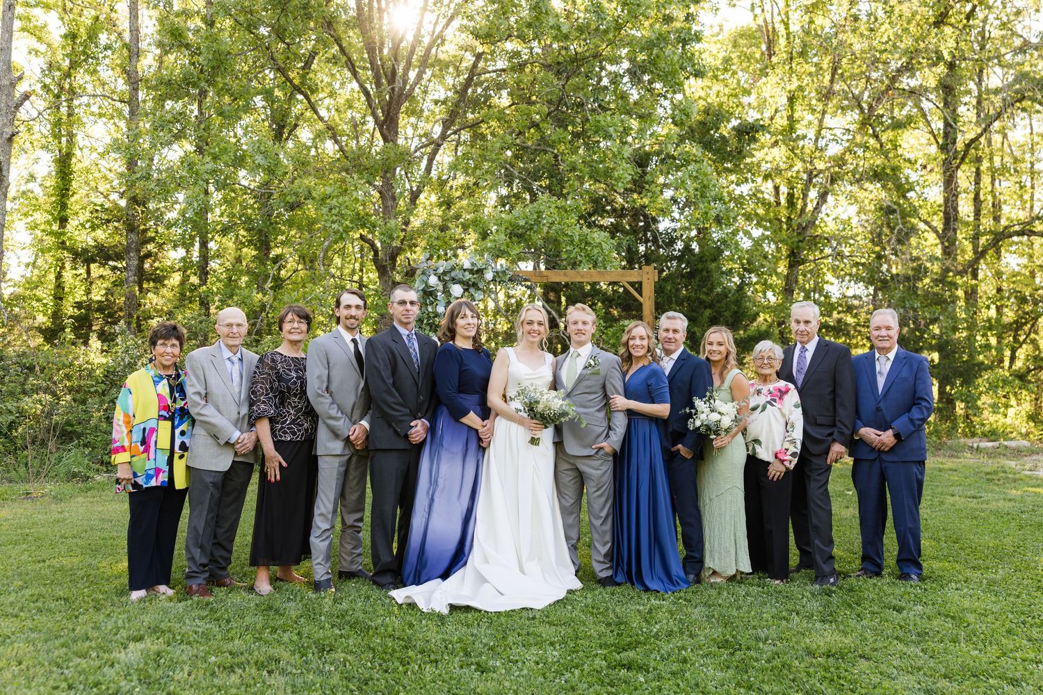 Wedding party poses outdoors in front of wooden arch, bride in white, bridesmaids in blue gowns, surrounded by lush green trees.