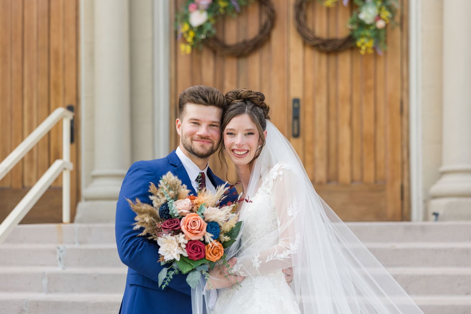 Smiling bride and groom pose together outside a church, bride holding a colorful bouquet of roses and dried flowers on their wedding day.