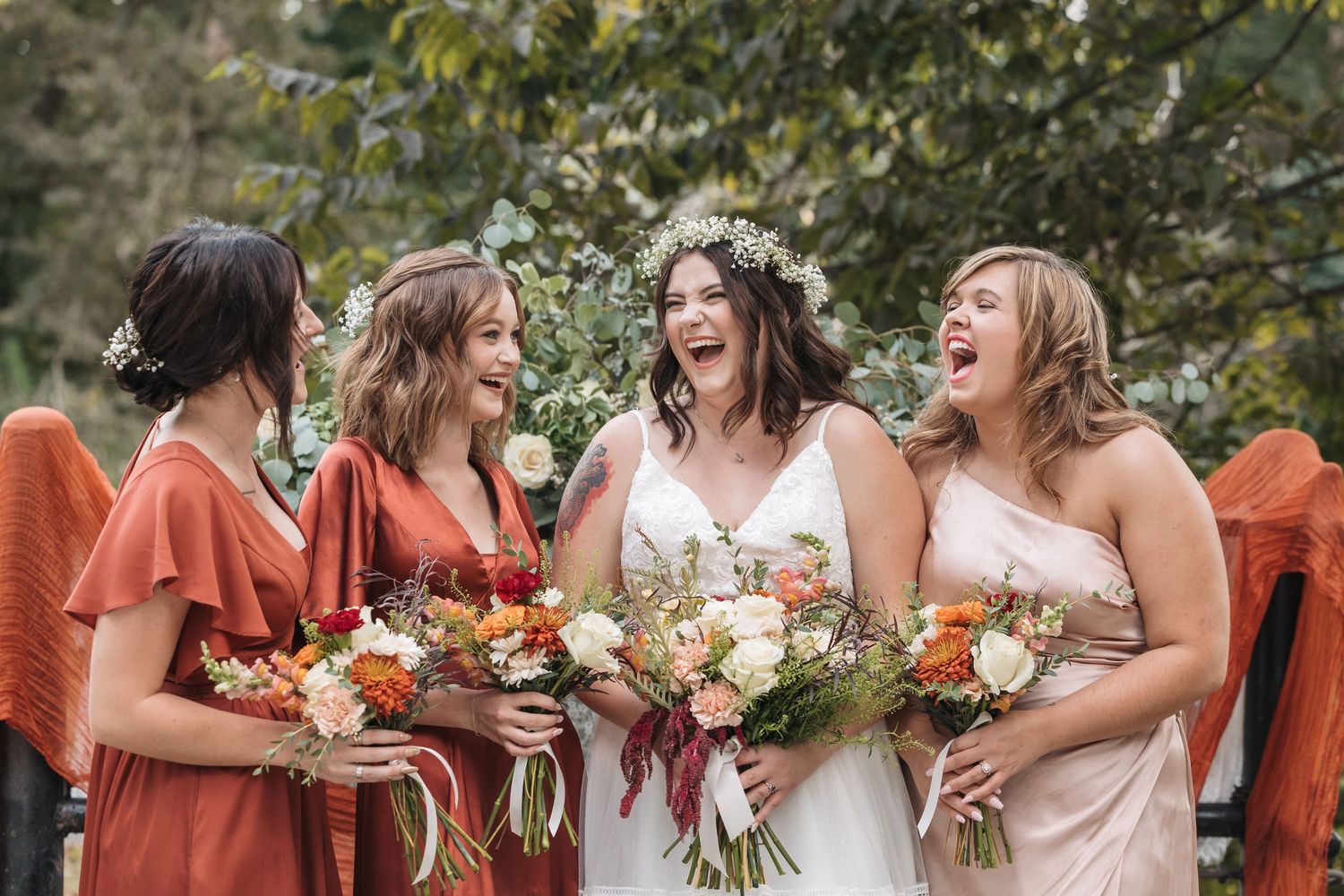 Bride and three bridesmaids in rust and blush dresses laughing together while holding colorful wedding bouquets outdoors.
