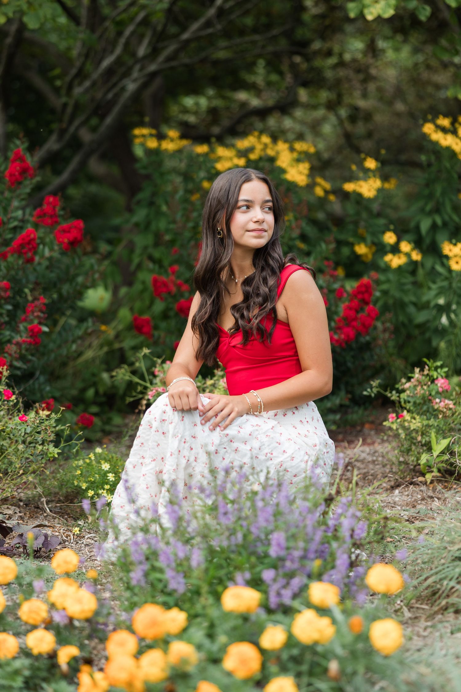 A portrait in a summer garden with vibrant red and yellow flowers, featuring a flowing white floral skirt and red top.