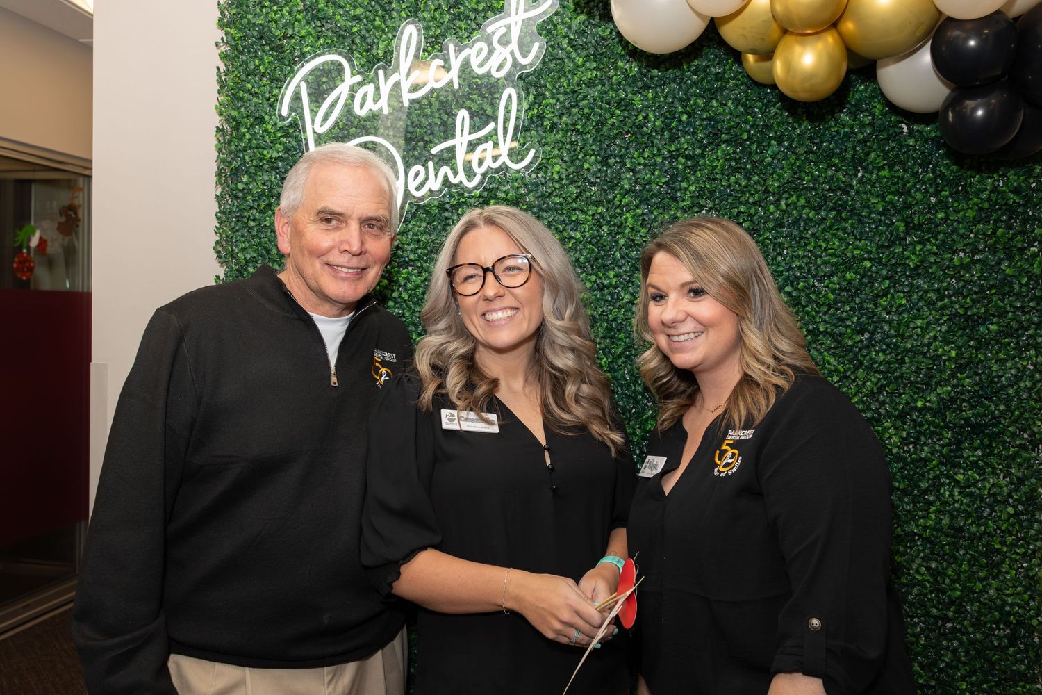 Group photo in front of green wall with gold balloon decorations at a dental office celebration.