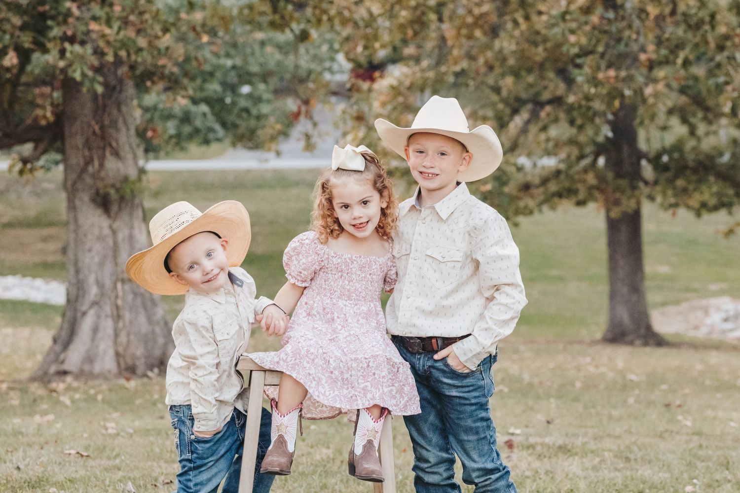 Three children in cowboy hats and western attire laugh together during an outdoor photoshoot.