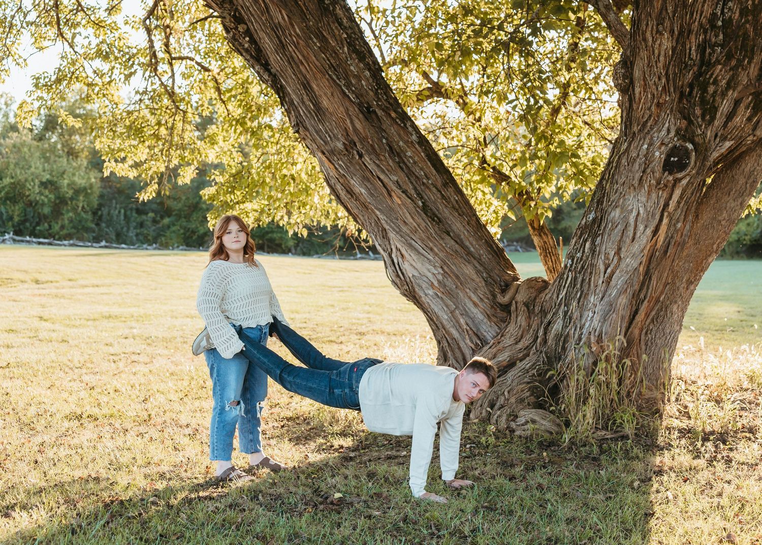 Two people playing and posing around a large tree trunk in golden afternoon light.