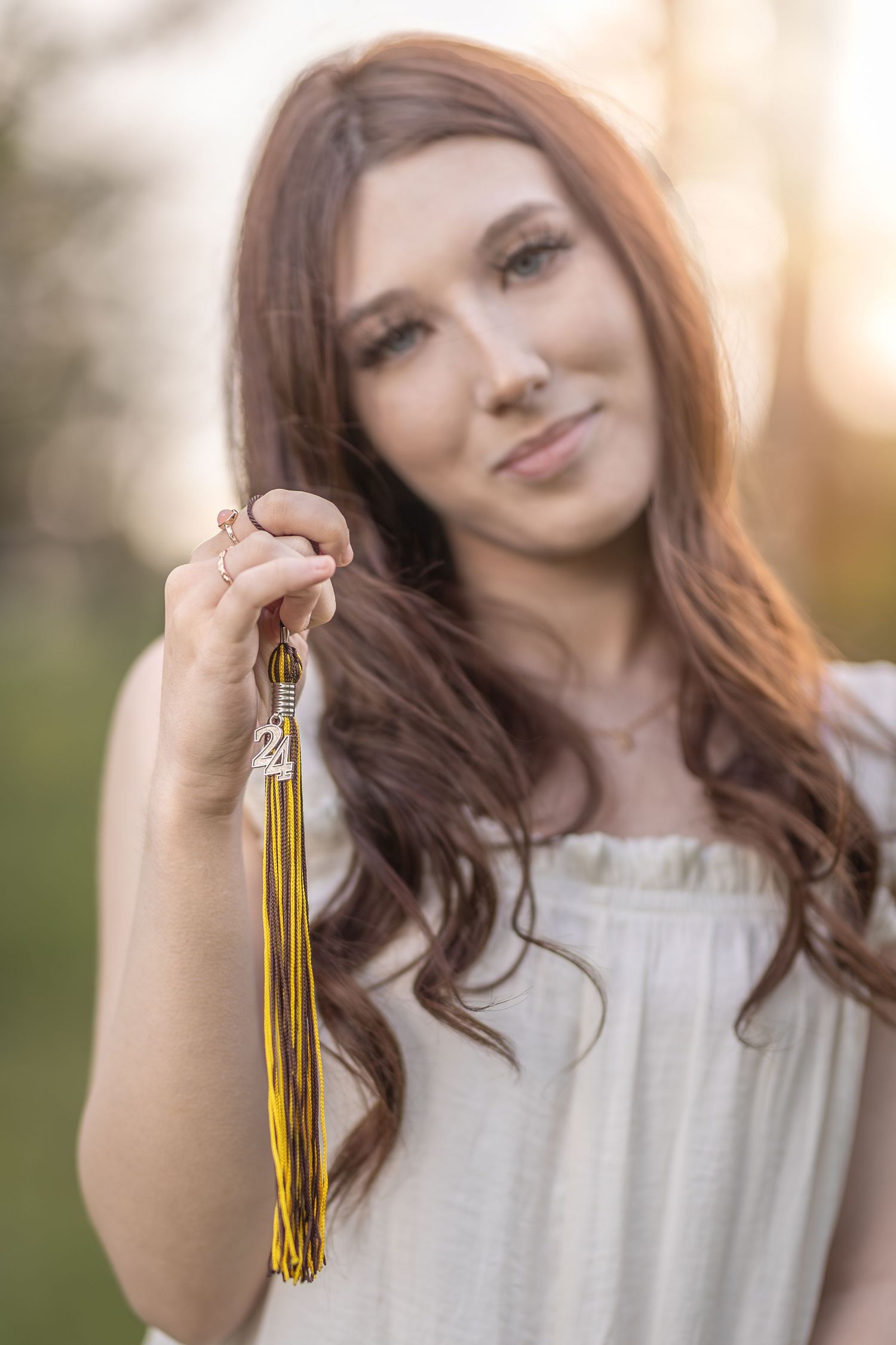 Someone holds gold graduation tassels while wearing white in a sunlit outdoor setting.