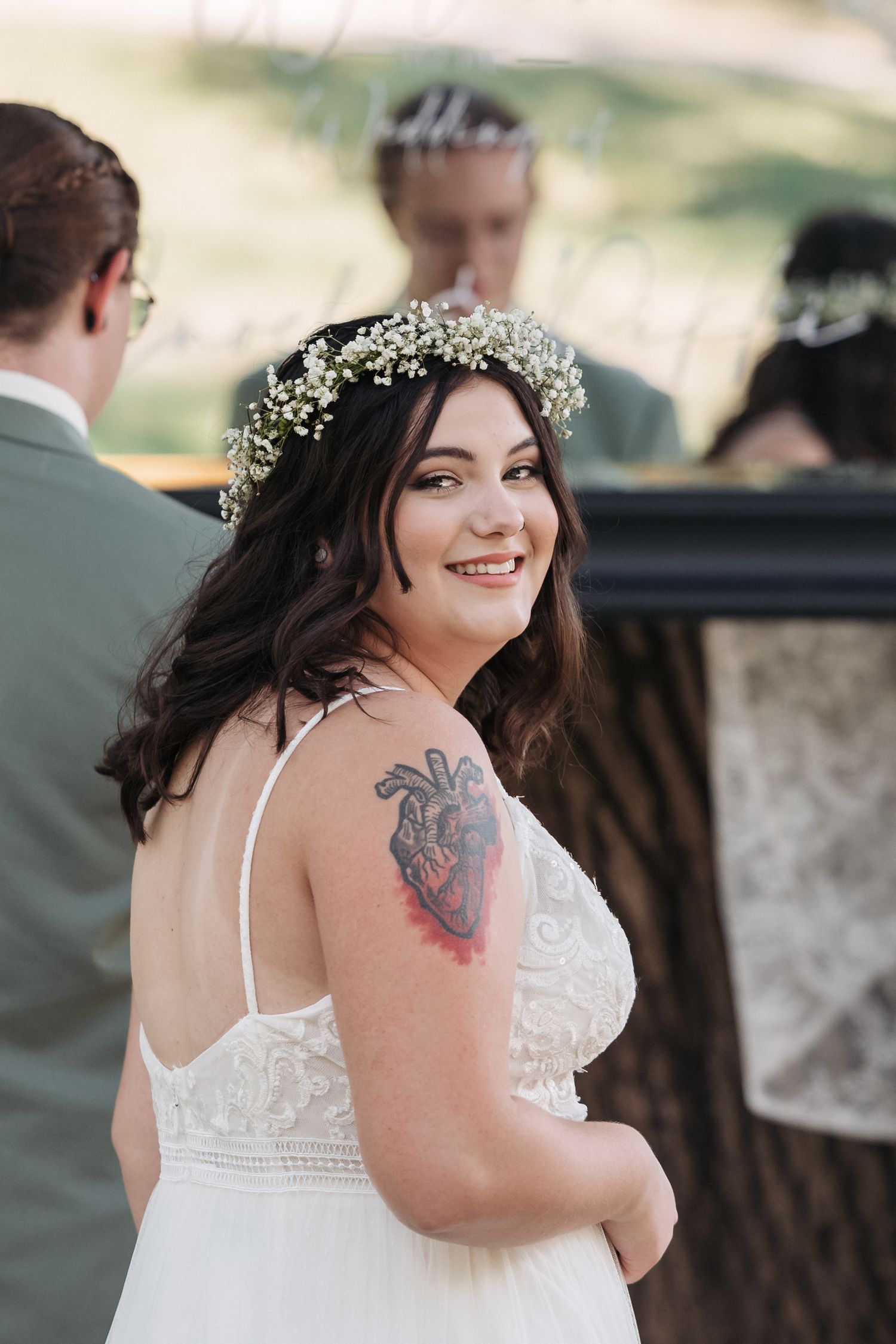 Bride wearing a lace dress and flower crown with visible tattoo smiling at wedding.