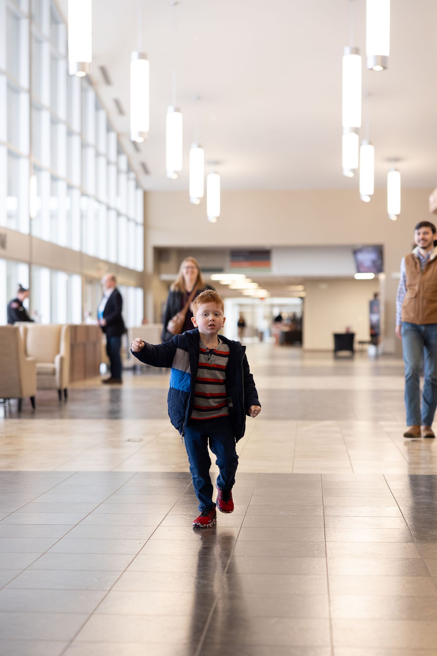 A boy running through a bright indoor hallway with high ceilings and hanging lights.