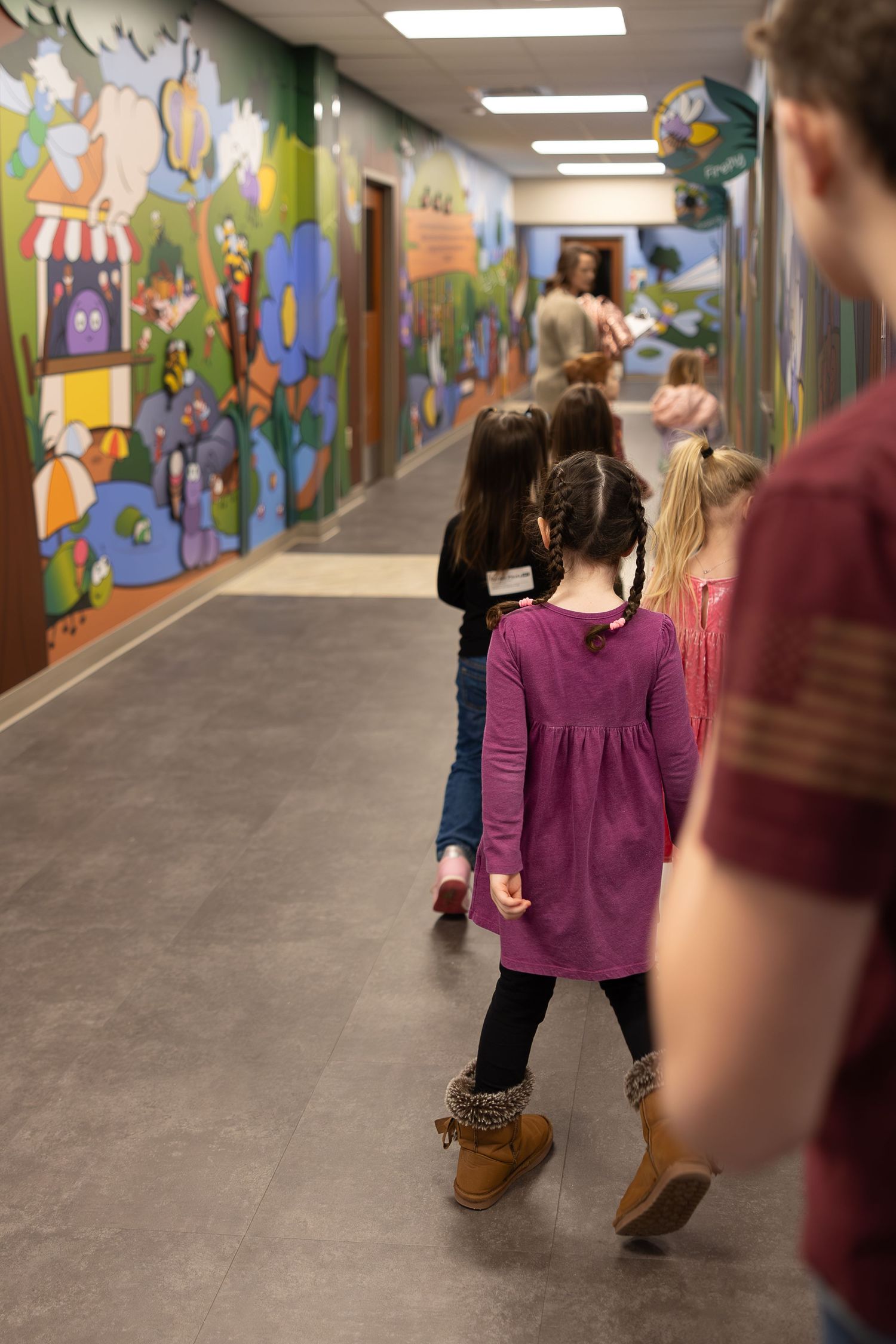 Children walking single file down hallway with vibrant cartoon murals on walls.