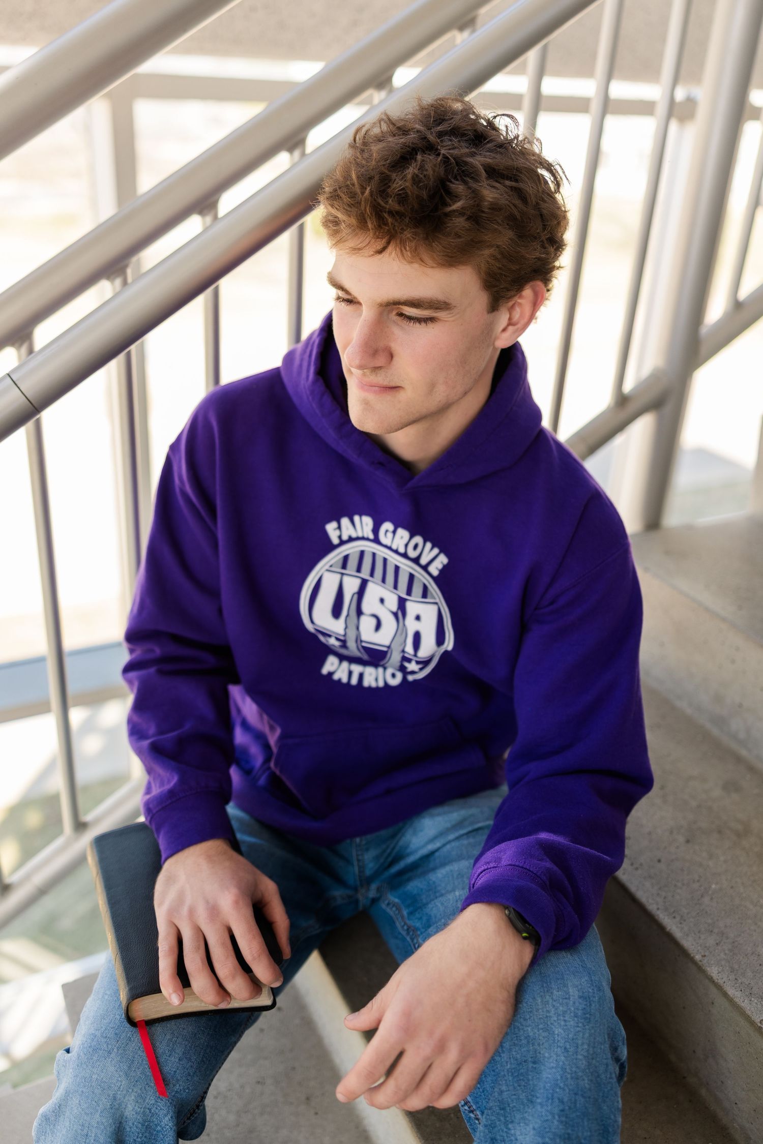 Someone wearing a purple USA Patriots athletic hoodie and jeans sits pensively on stairs in a well-lit space.