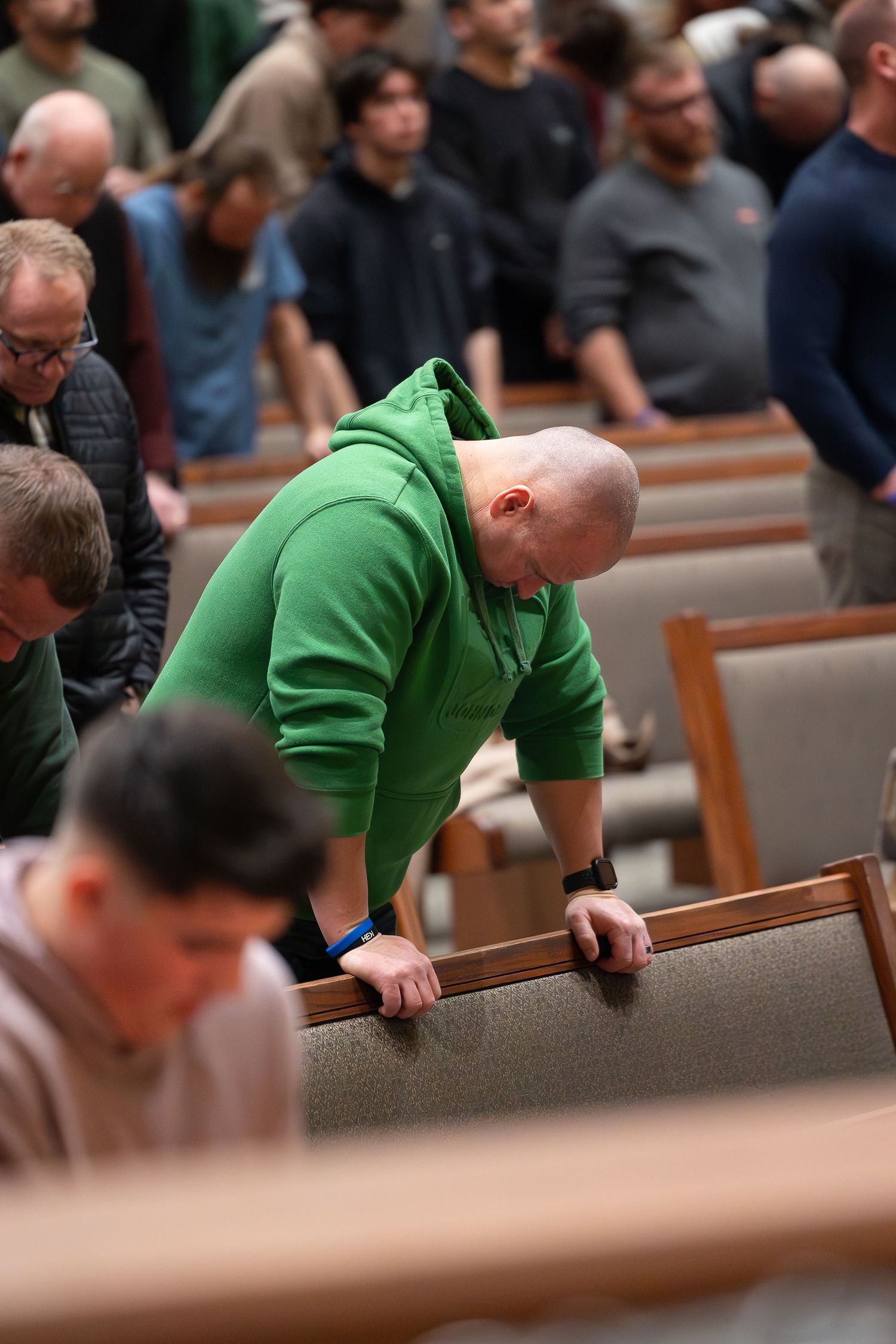 A person wearing a bright green hoodie leans over a wooden church pew in a crowded congregation.
