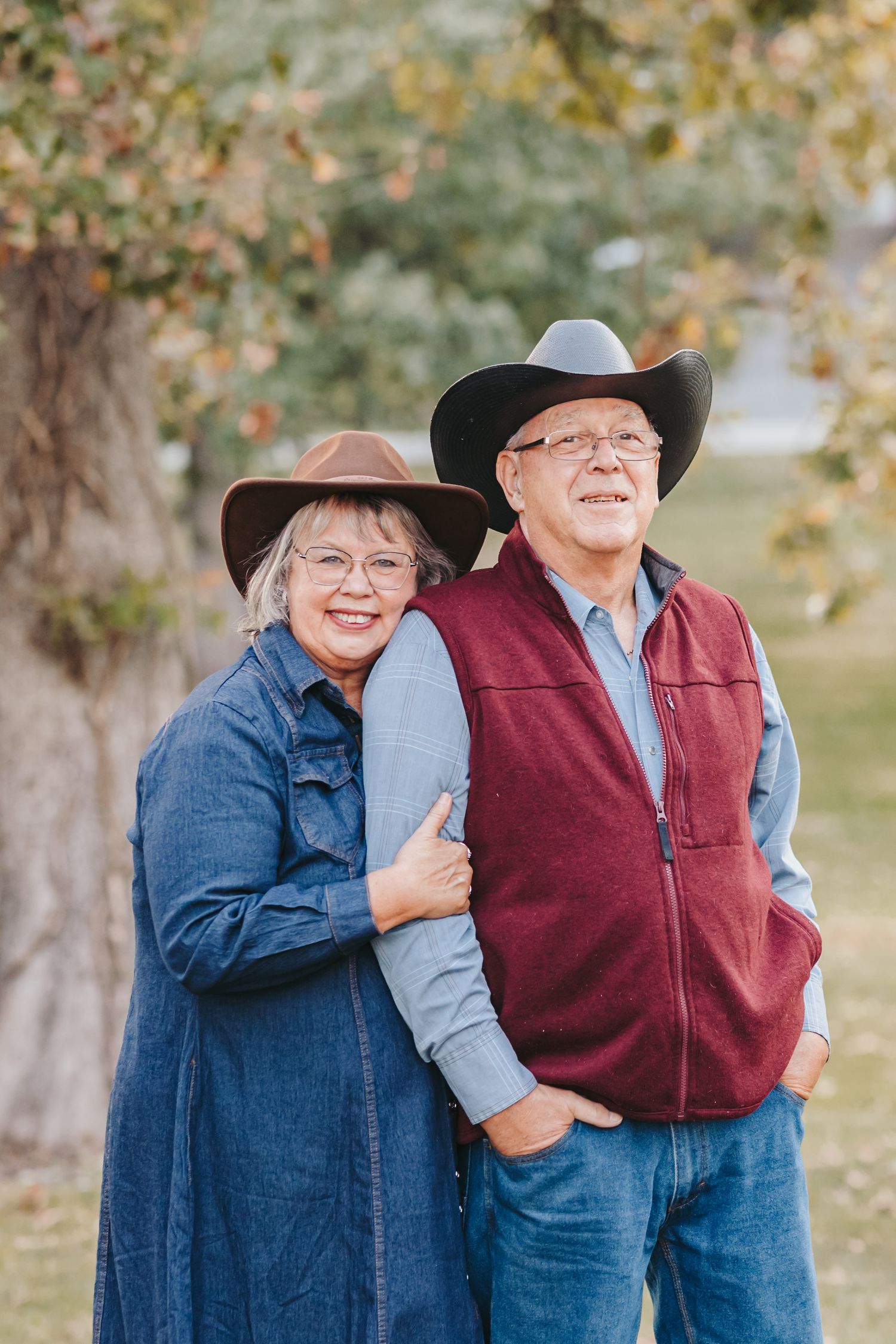 Couple in western attire and cowboy hats smile together outdoors.