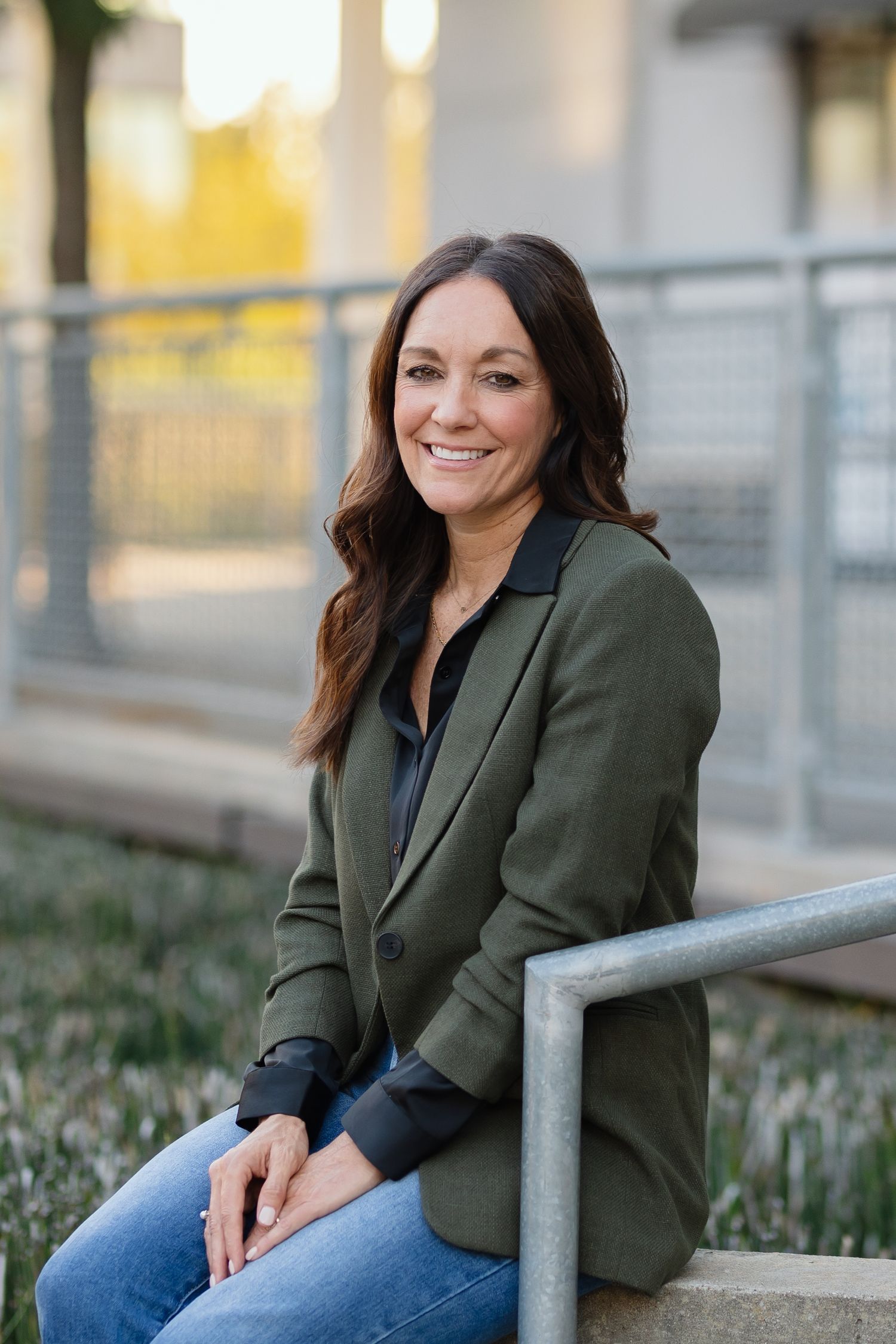 Portrait of person in olive blazer and jeans smiling while sitting outdoors.