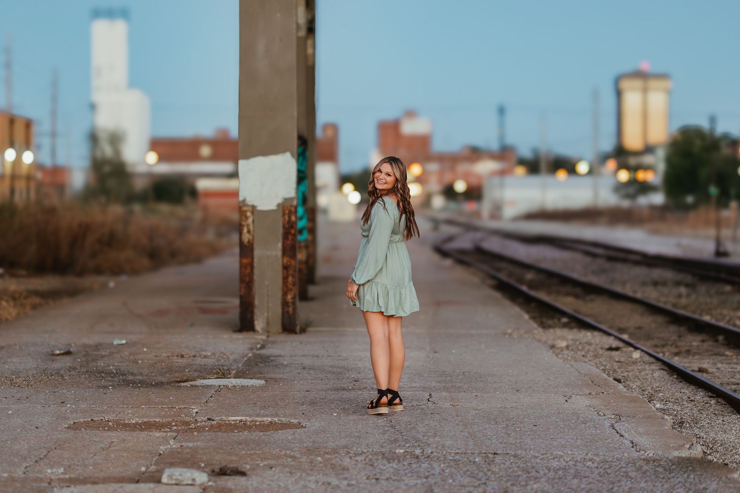 Person in flowy mint dress stands on train tracks in urban setting.
