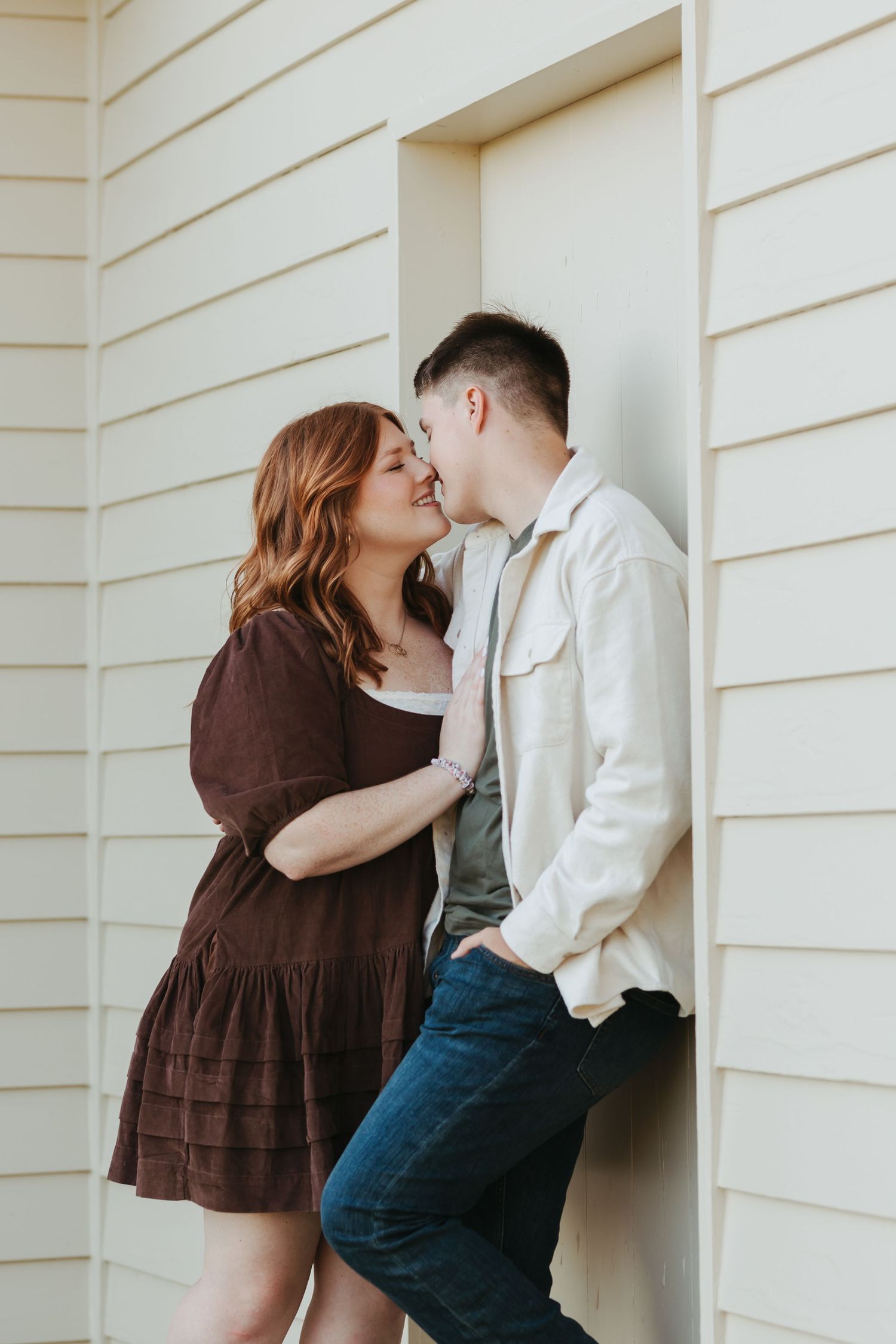 Couple shares romantic kiss against white wall, dressed in brown dress and light denim outfit.