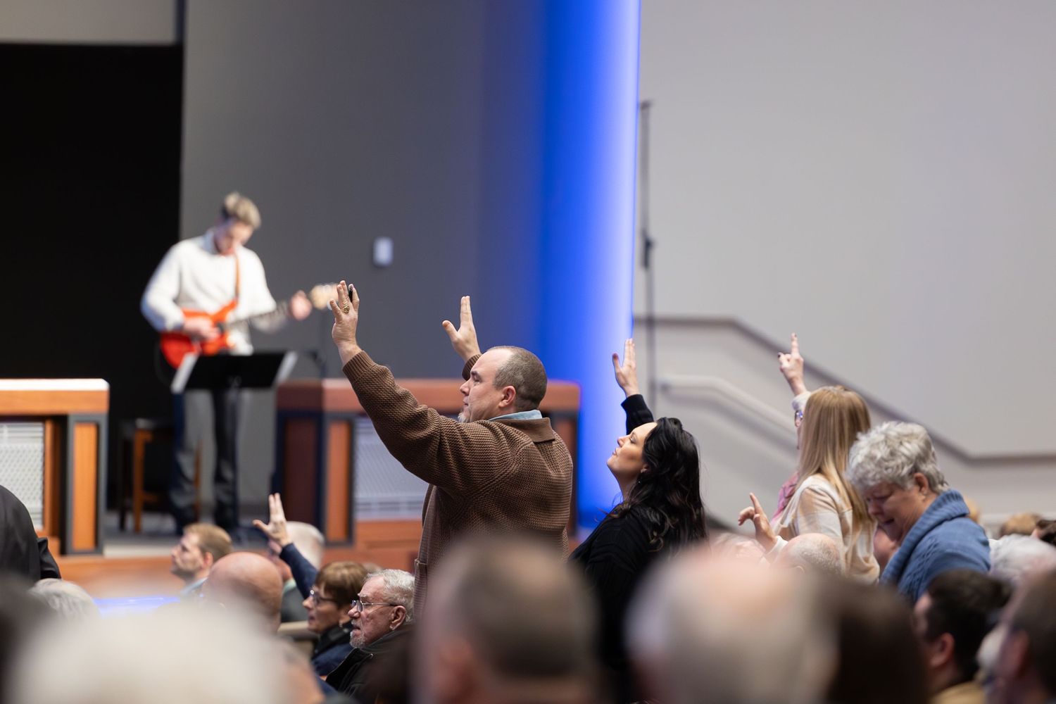 Audience members raising hands during worship service with musicians performing on stage.