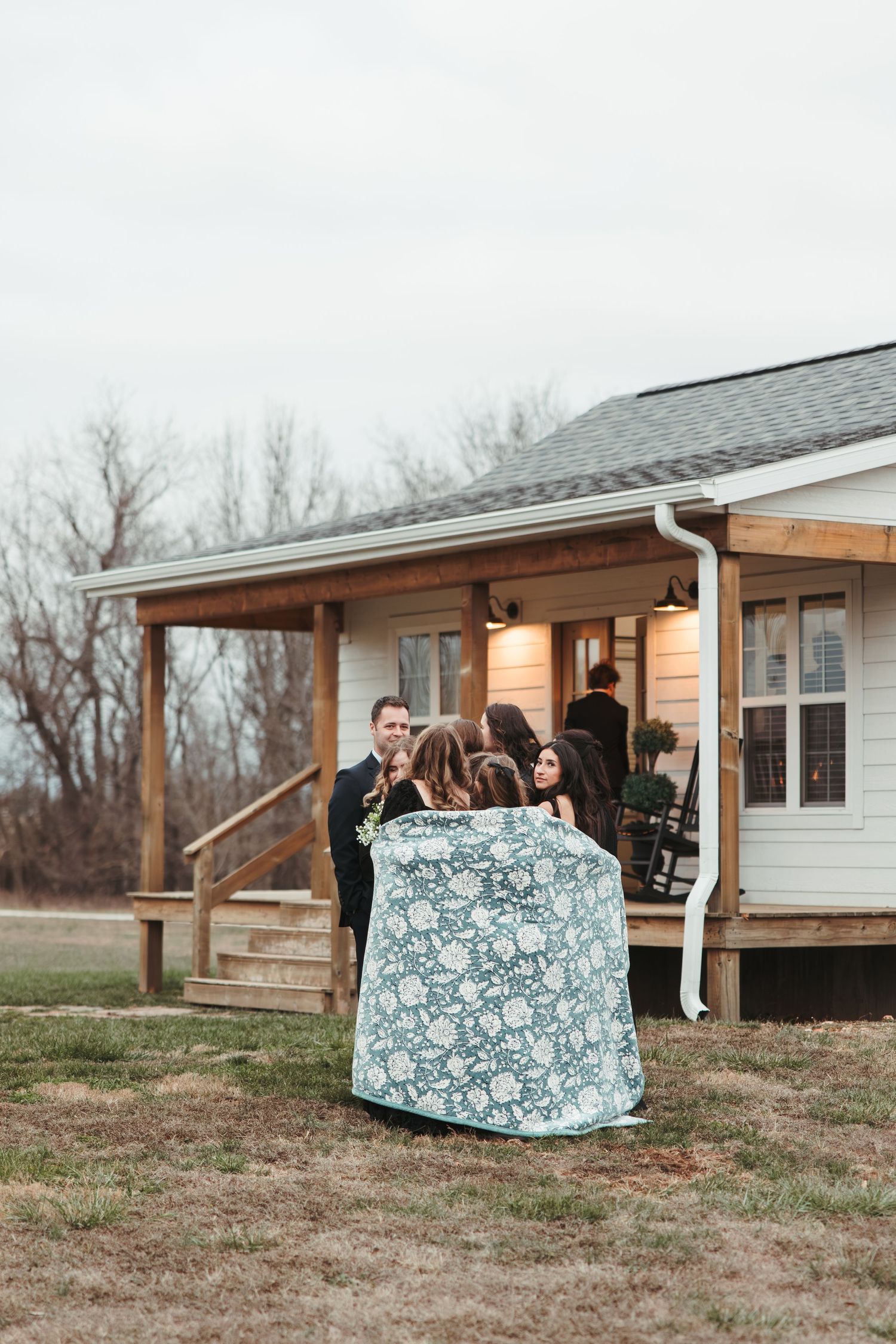 Wedding party gathers on the porch of a rustic white farmhouse.