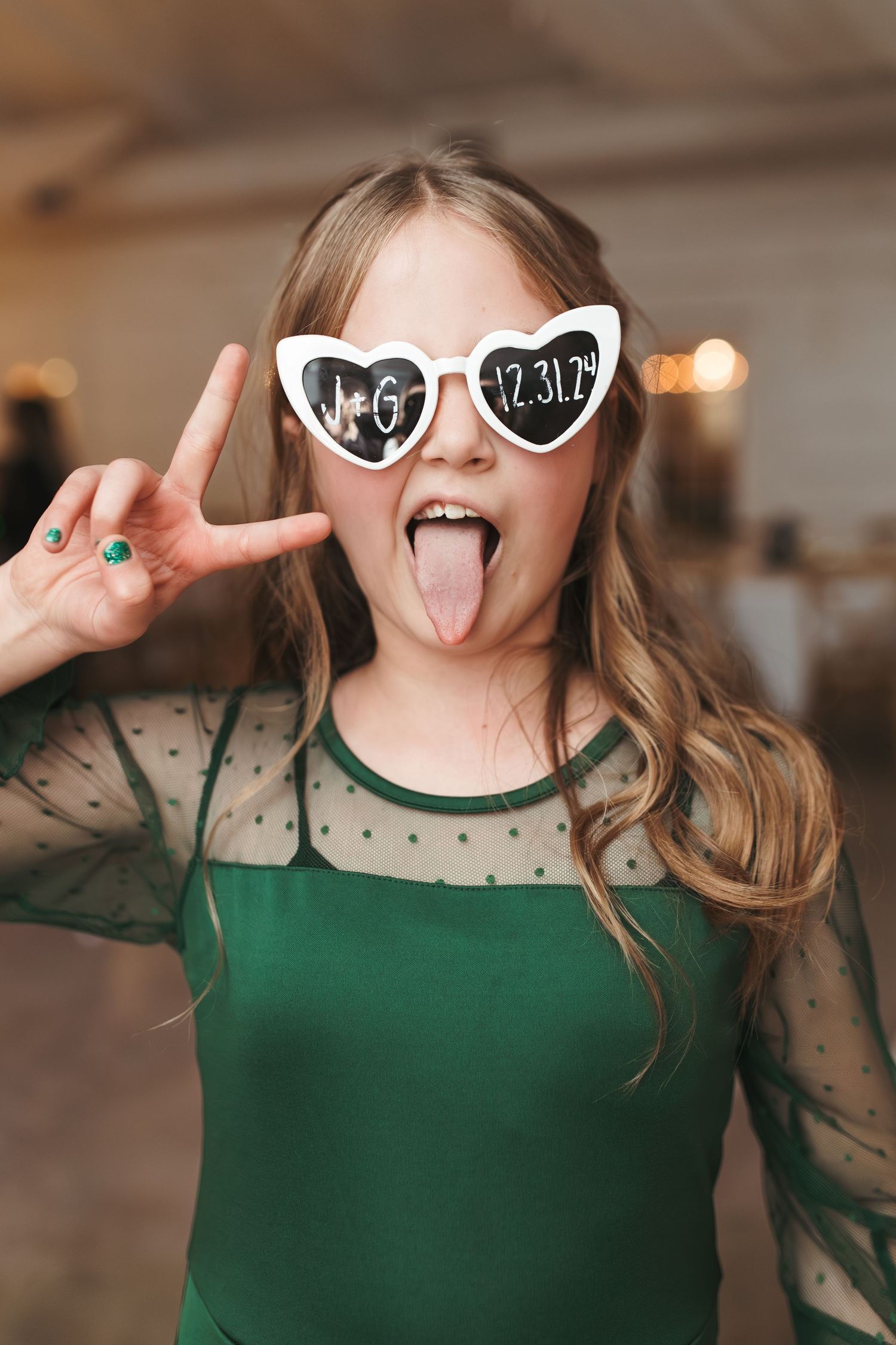Person wearing heart-shaped sunglasses and green dress makes peace sign at party.