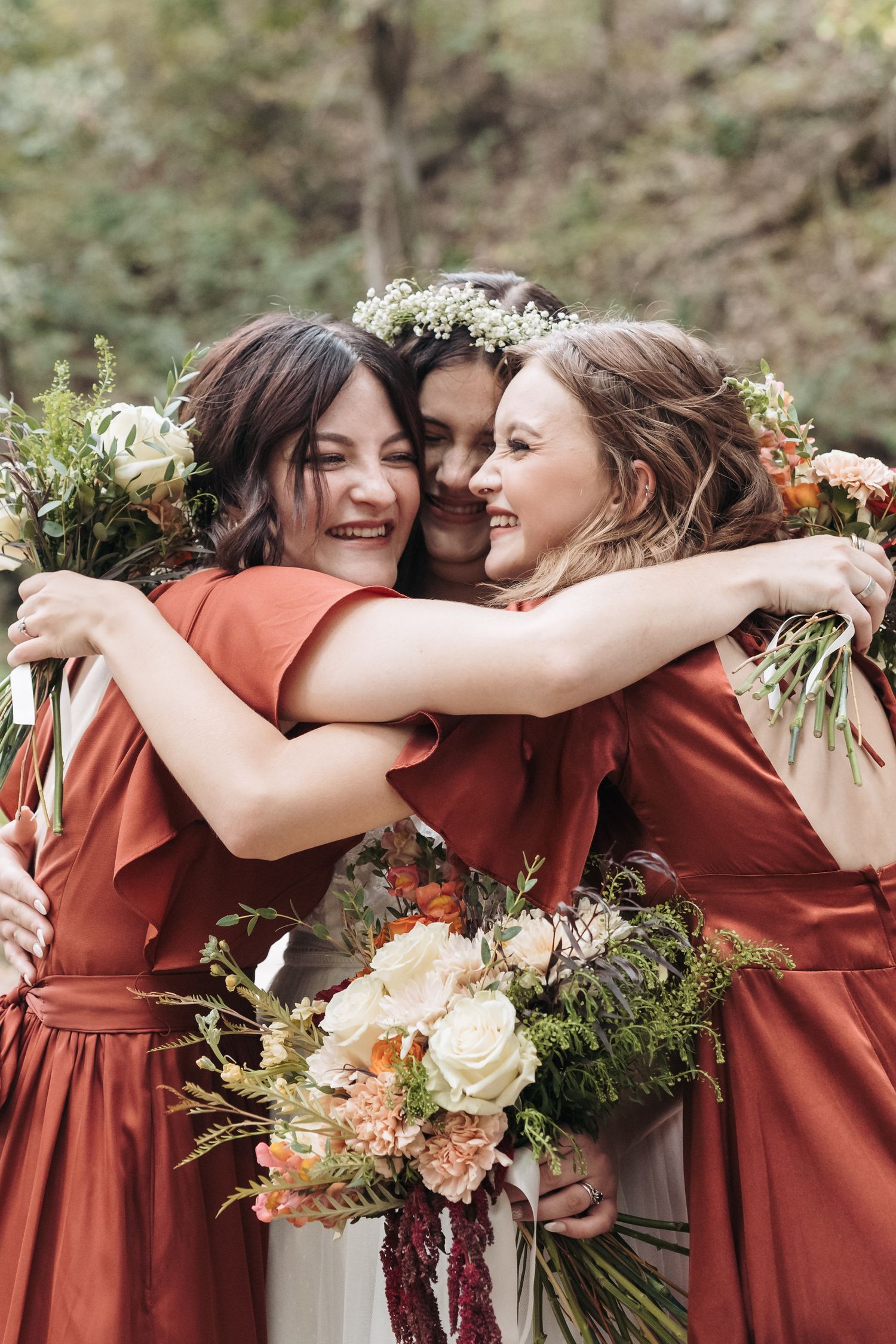 Three people in coral dresses embrace while holding floral bouquets.