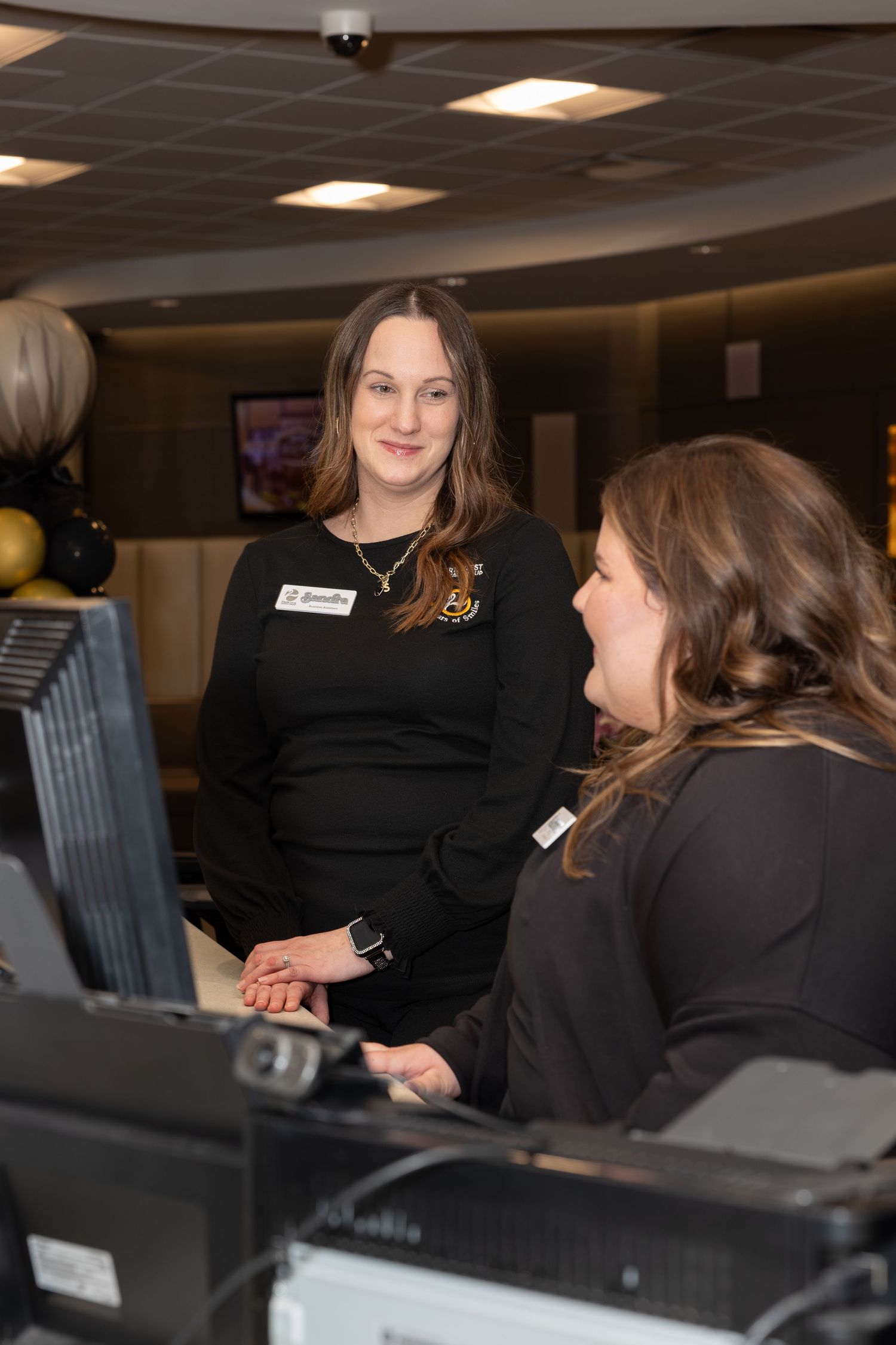 Two staff members in dark uniforms interact at computer workstation under office lighting.