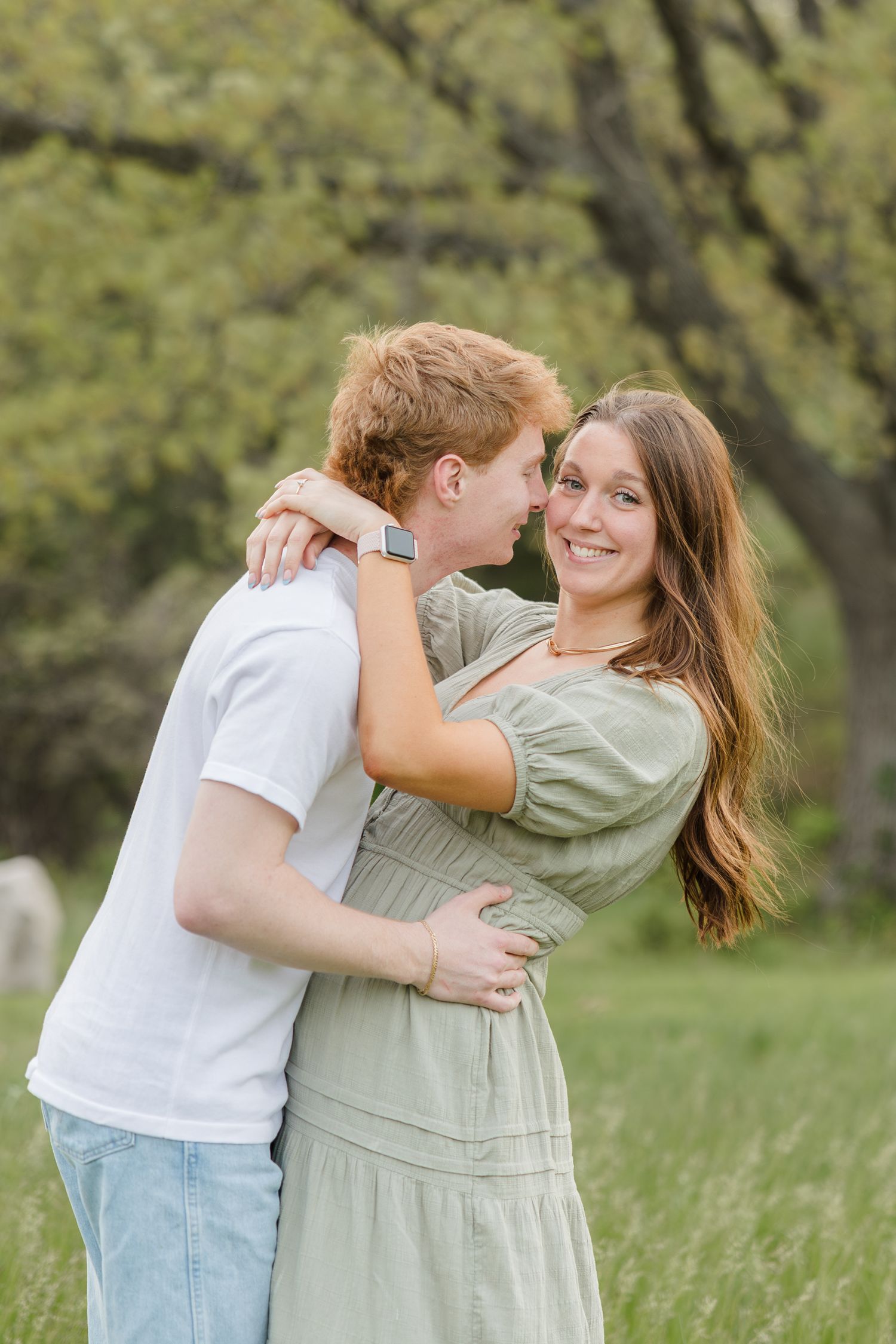 A romantic couple embraces in a grassy field with trees in the background on a spring afternoon.