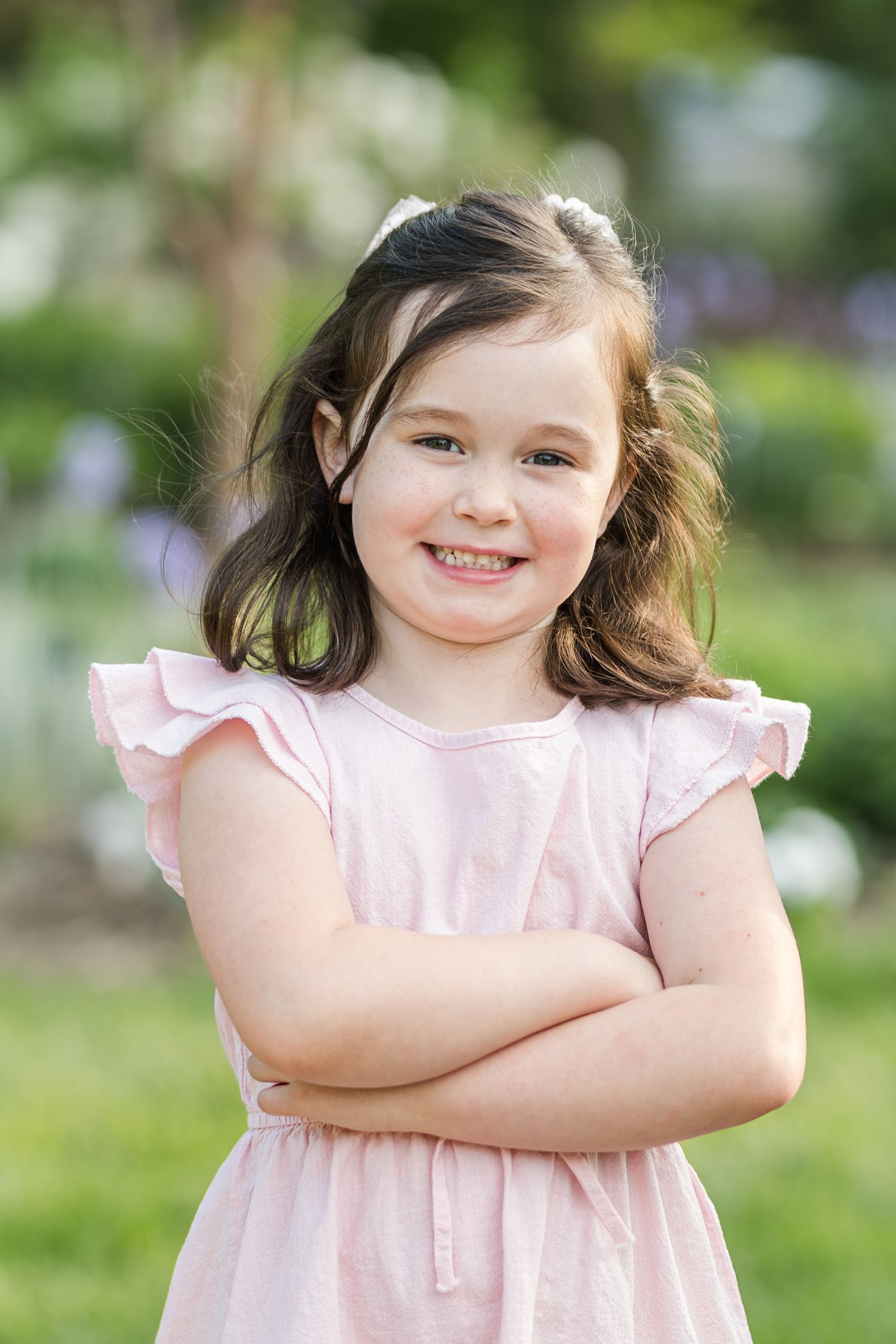 Young child wearing a light pink ruffled dress stands with arms crossed in garden setting.