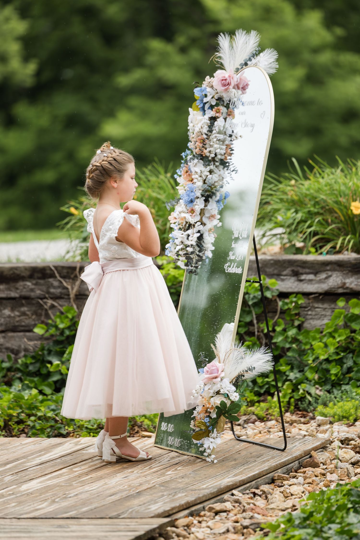 Young flower girl in pink dress admires floral wedding welcome mirror sign outdoors.