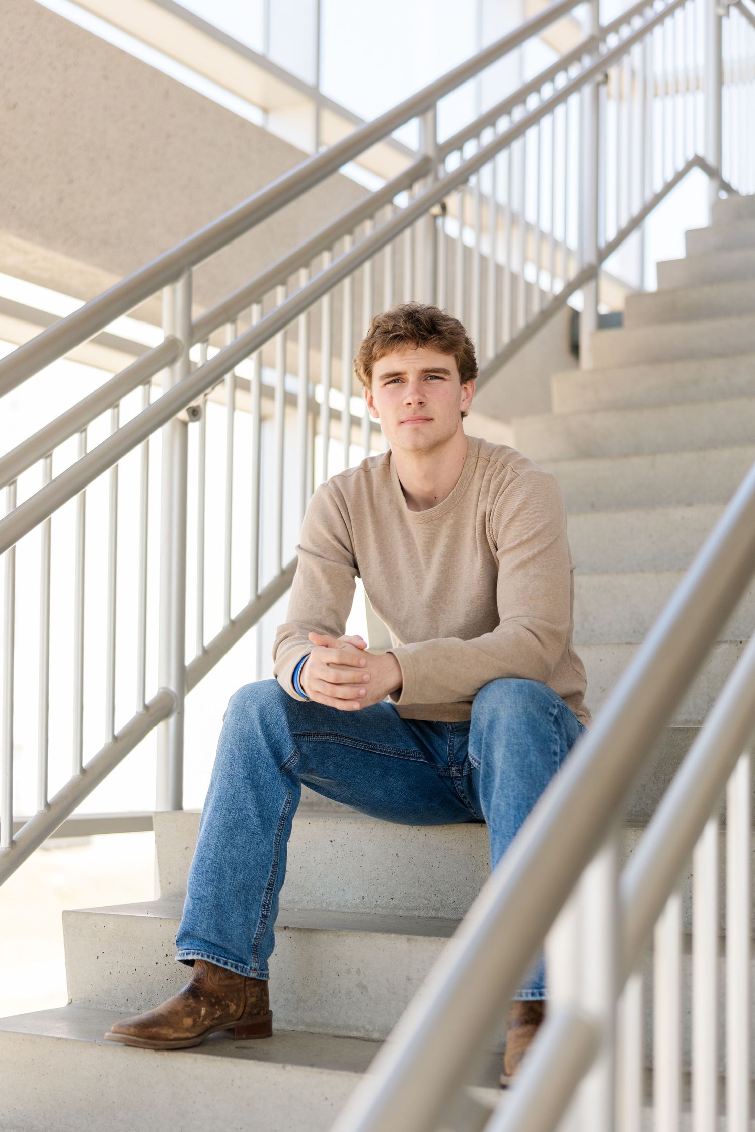 Person in neutral sweater and jeans sits thoughtfully on concrete stairs with metal railings in natural light.