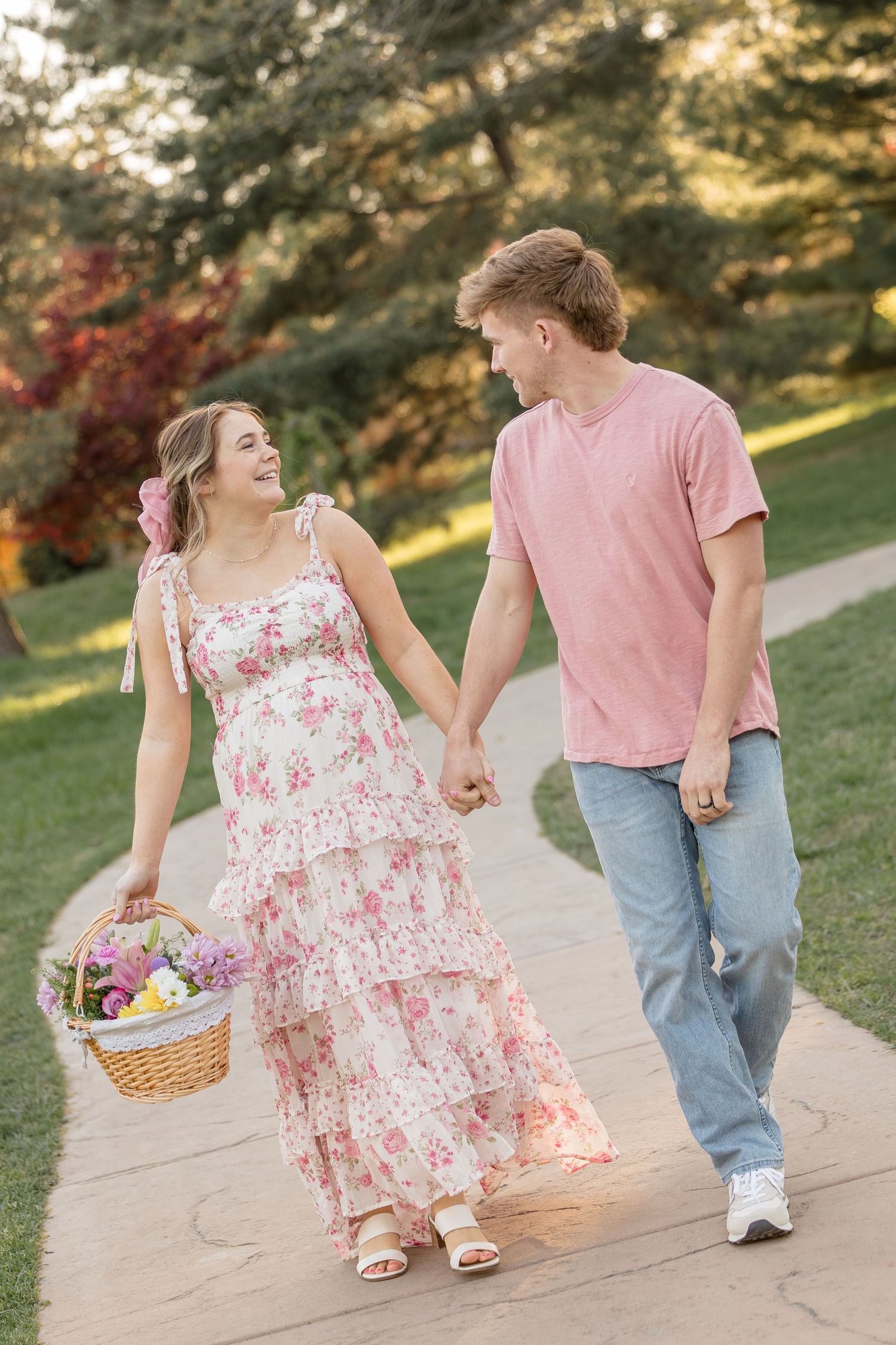 A couple walks together on a garden path, with her holding a basket while wearing a floral dress.