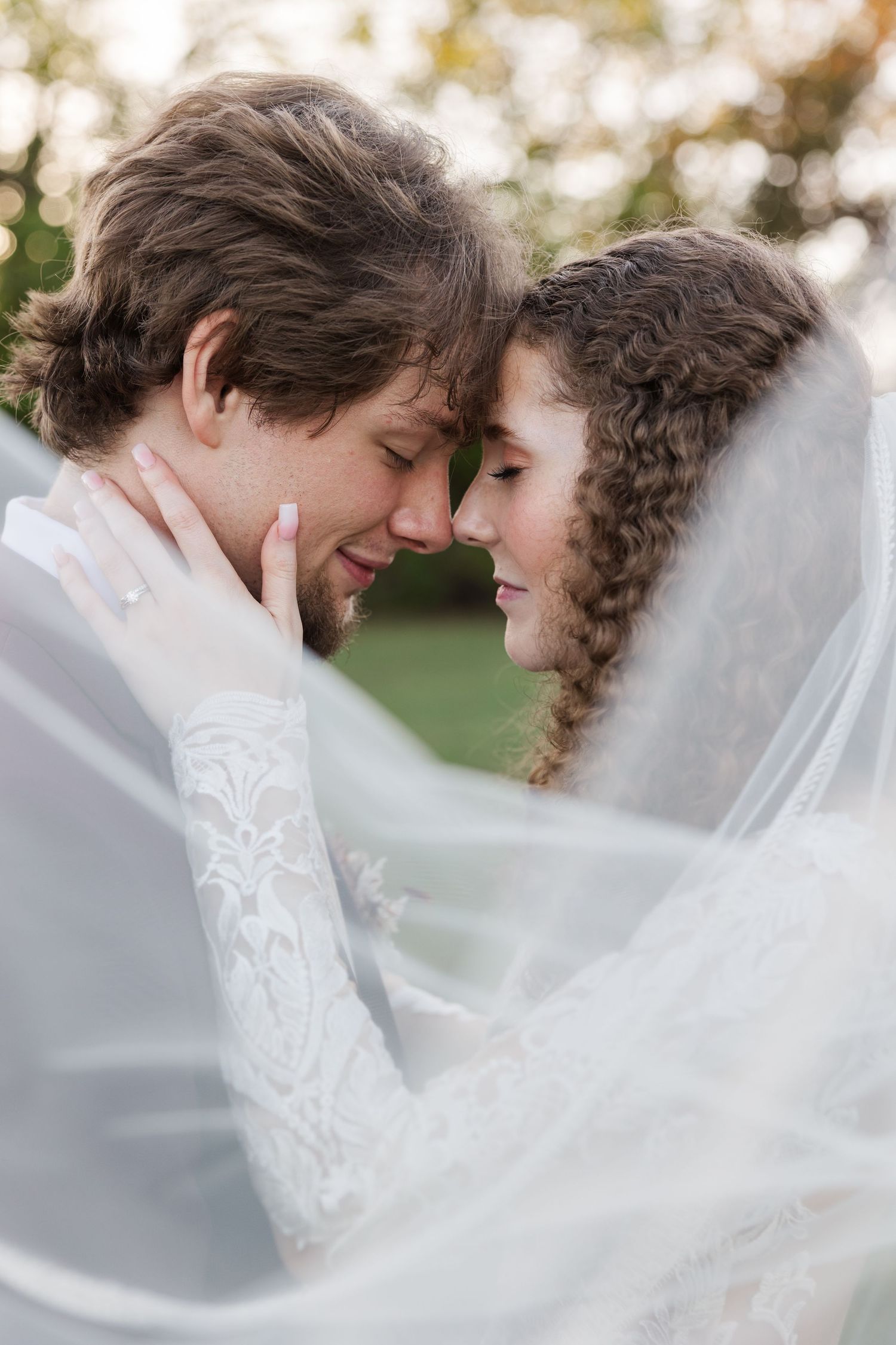 A romantic wedding couple shares an intimate moment under a flowing white veil in a dreamy outdoor setting.