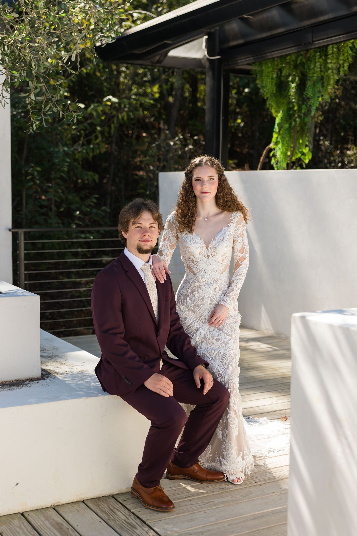 Bride in lace gown and groom in burgundy suit pose together on a sunlit outdoor venue deck.