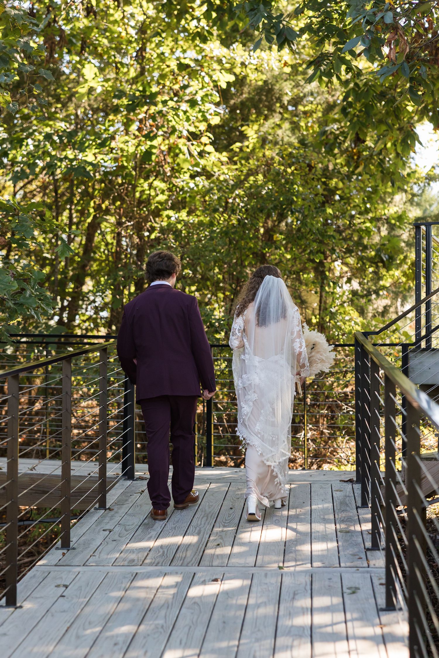 Bride and groom walking hand in hand across a wooden bridge surrounded by lush green trees on their wedding day.