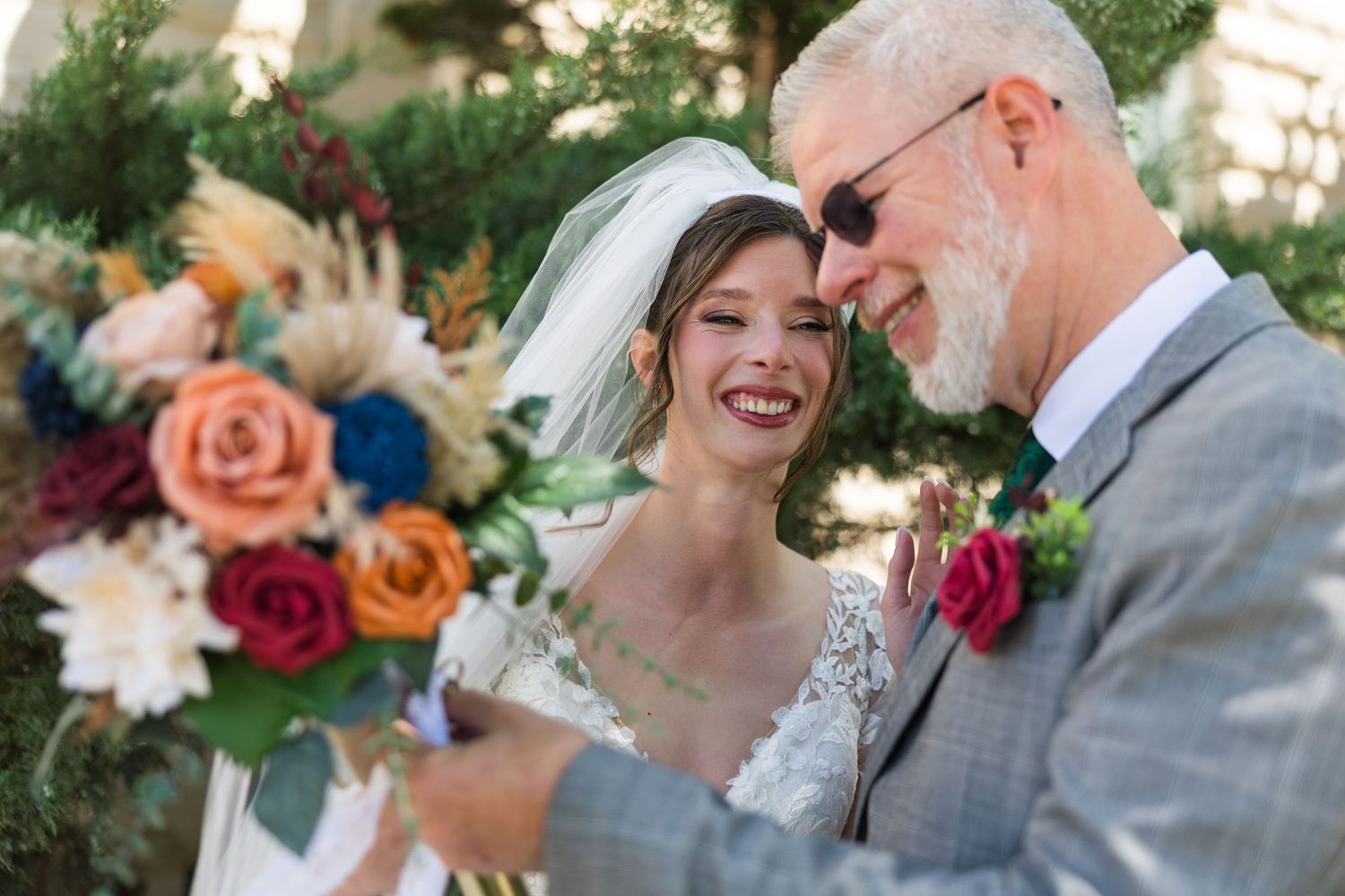 A smiling bride in a lace gown and veil shares a joyful moment with an older man holding a colorful bouquet.