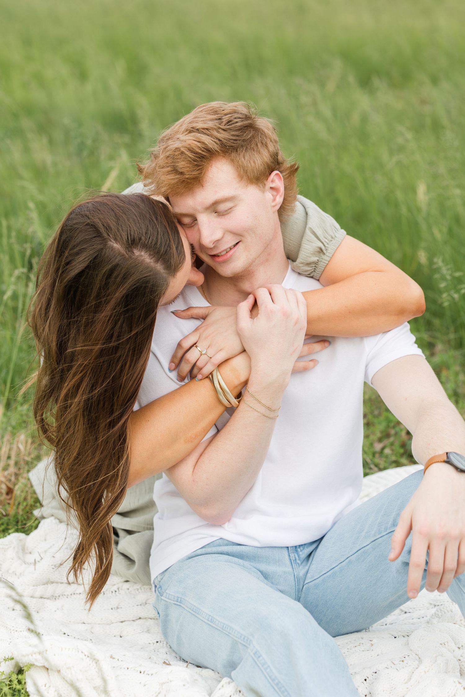 Romantic couple sits on blanket in tall grass field sharing intimate moment together on sunny day.