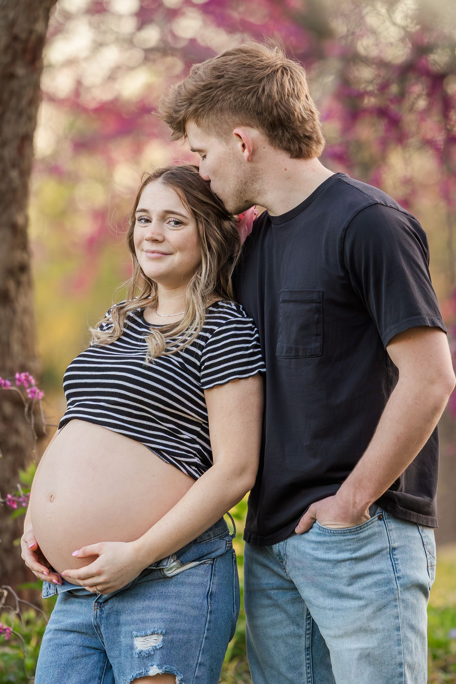 Expecting couple in casual clothing share tender moments while posing together outdoors with spring blossoms in background.